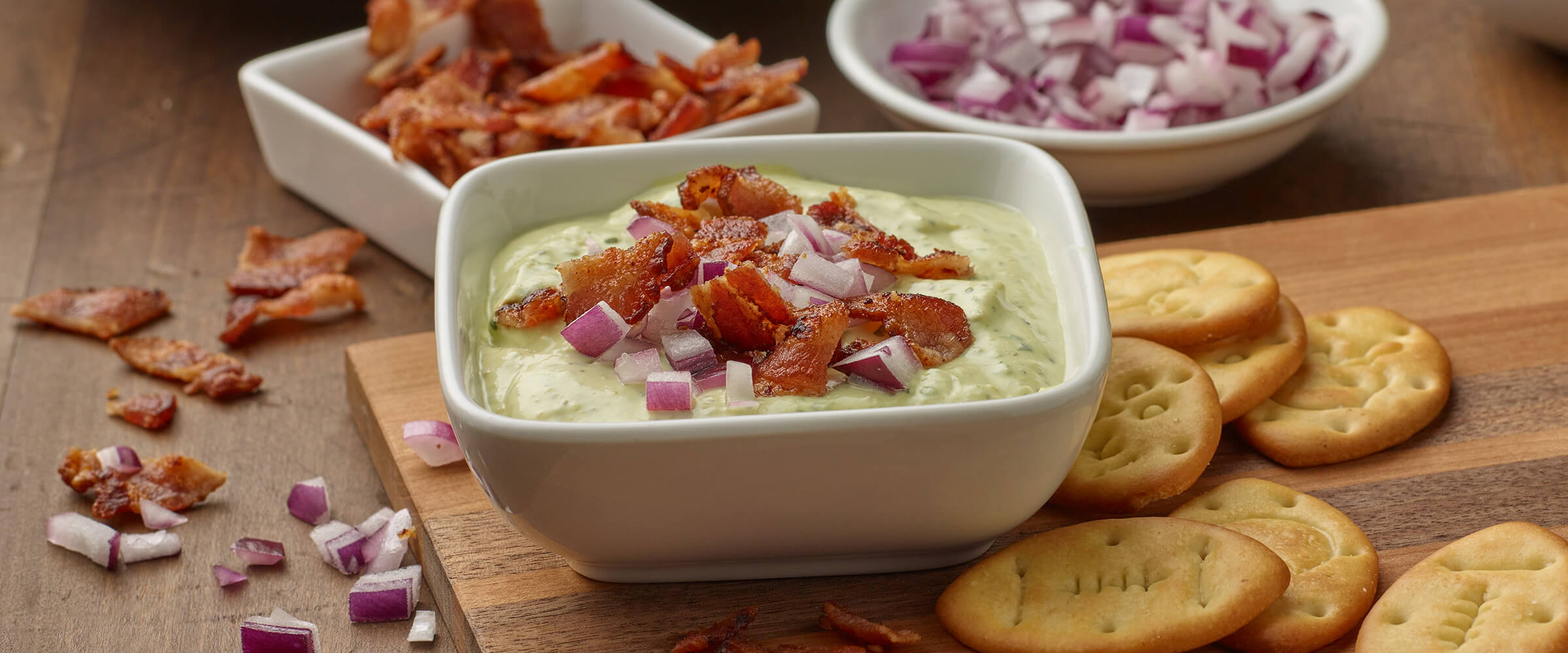 Creamy Avocado Jalapeno Dip topped with bacon and red onions in white bowl with crackers on the side