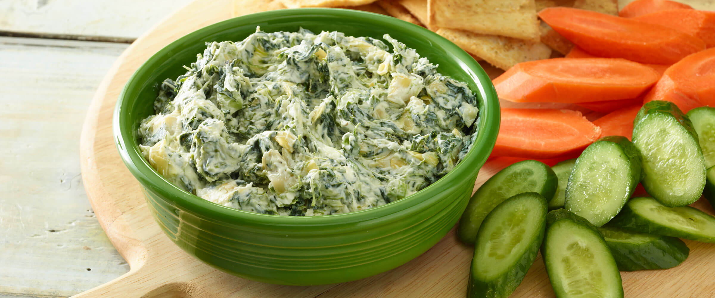 Spinach and Artichoke dip in green bowl with cucumber and carrot dippers