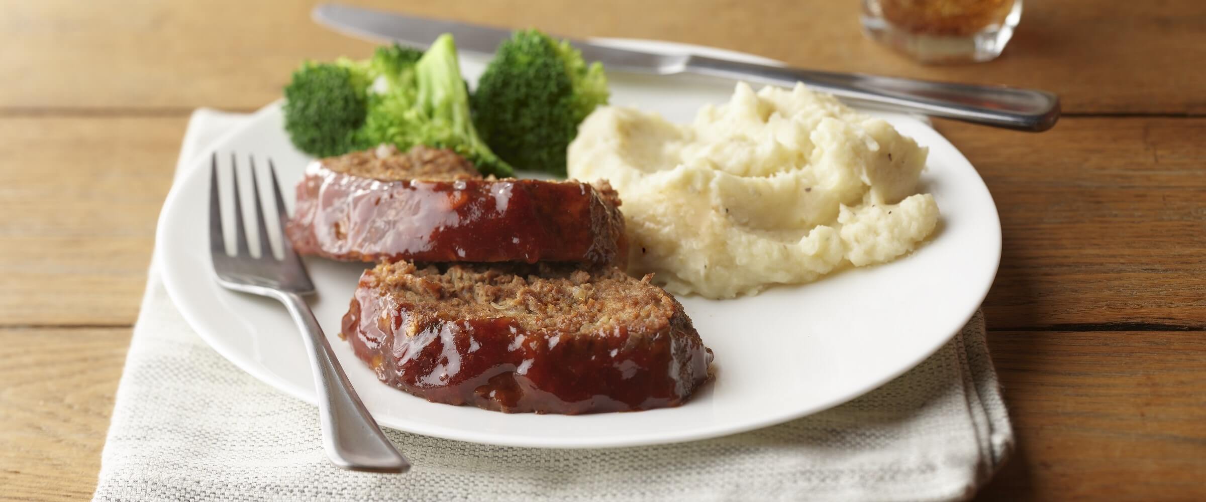 Glazed meatloaf with mashed potatoes and broccoli on a white plate with silverware