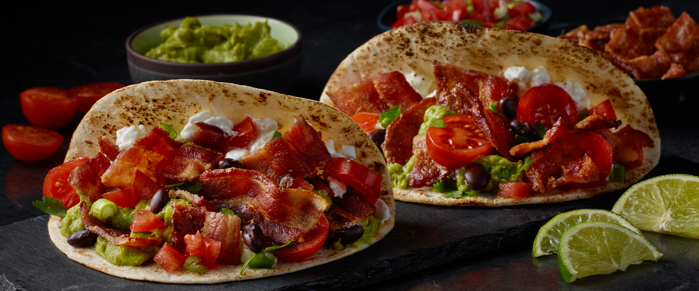 Two bacon and avocado breakfast tacos on a black serving board. Two lime wedges are on the right side of the tacos.