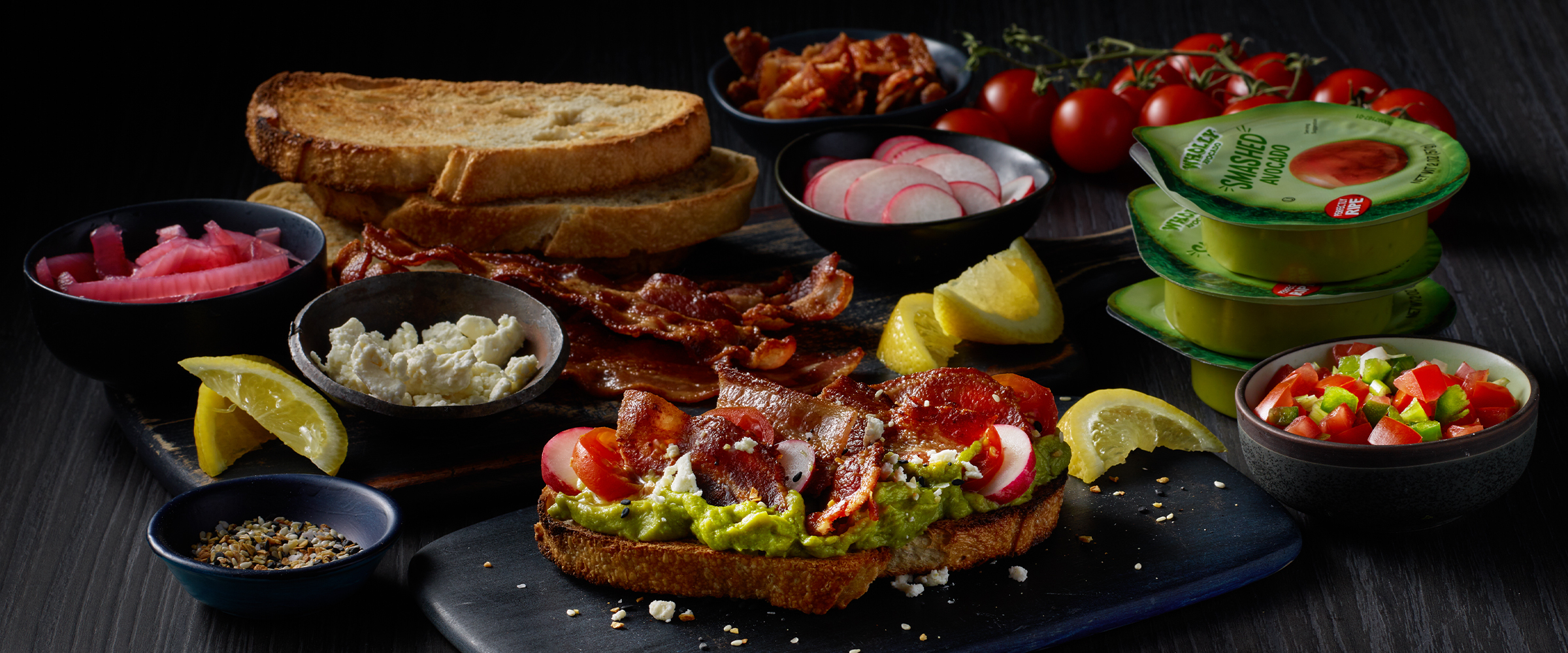 Bacon and avocado toast on a black serving board. Around the board are several toppings, including sliced and toasted bread, lime wedges, pickled onion and pico de gallo.