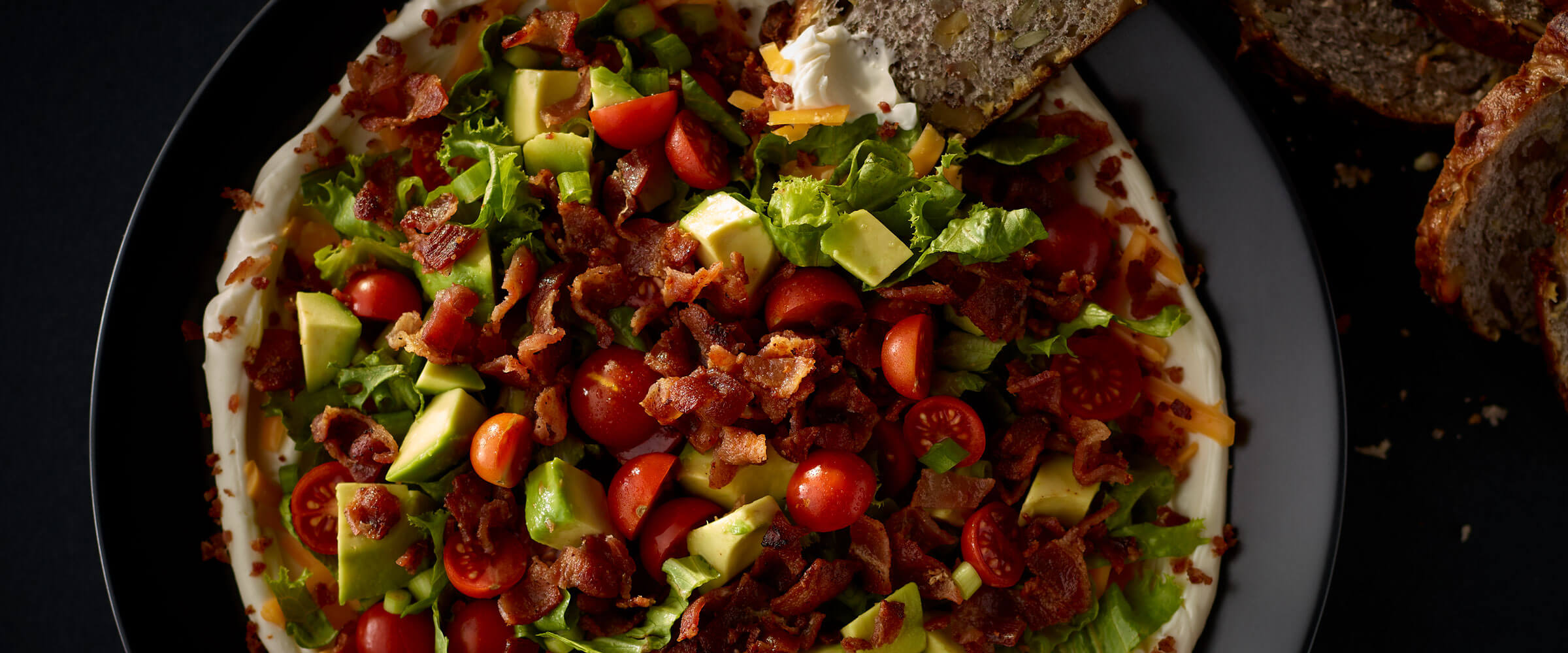 BLT Dip topped with avocado on black plate with bread for dipping