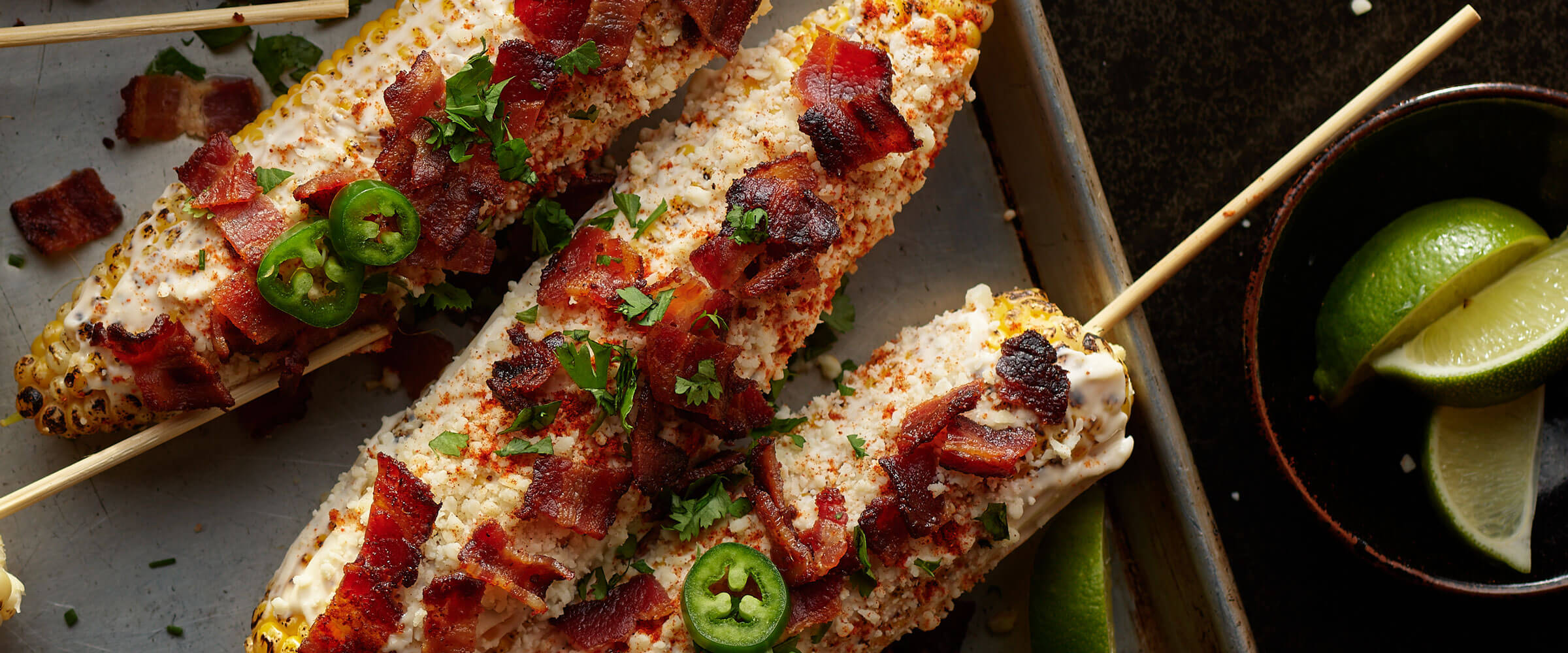 Jalapeno Bacon Grilled Elote corn on skewers with side of lime wedges
