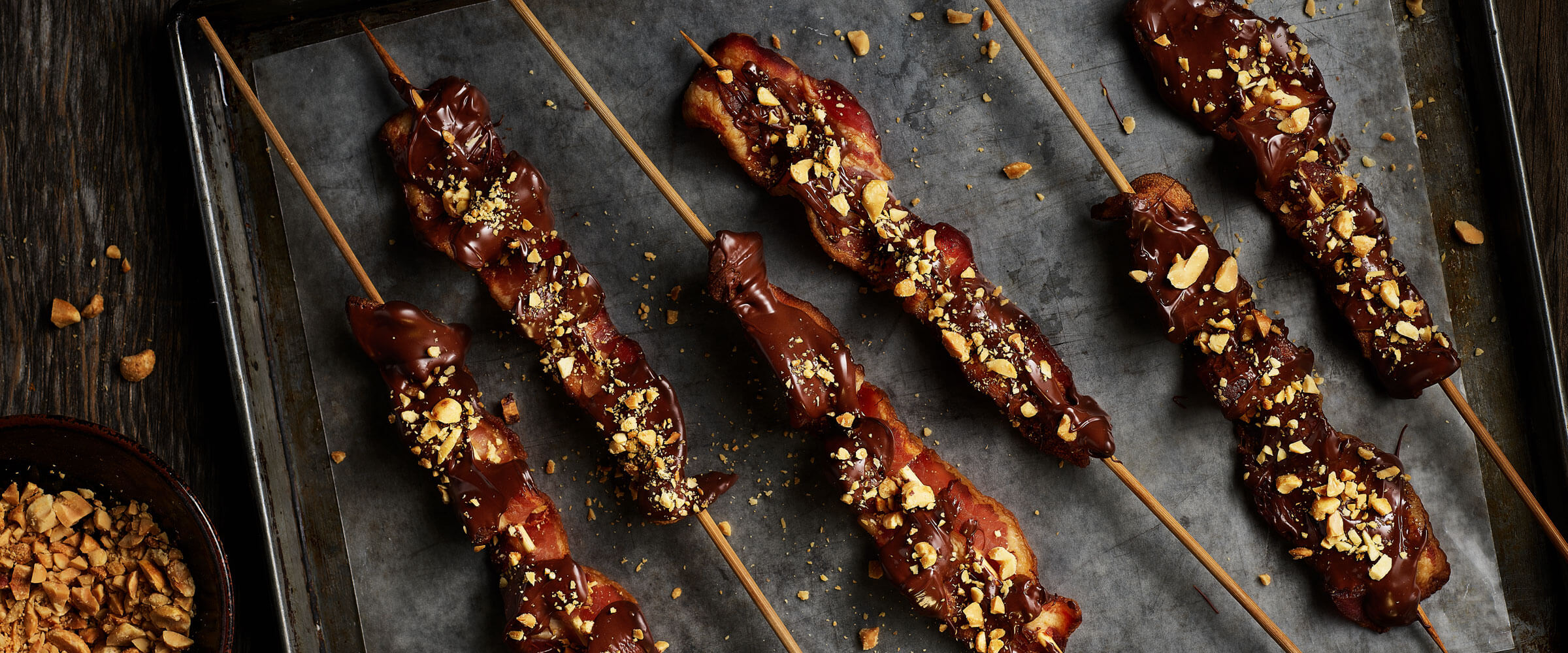 Chocolate-Covered Bacon on skewers topped with nuts on sheetpan