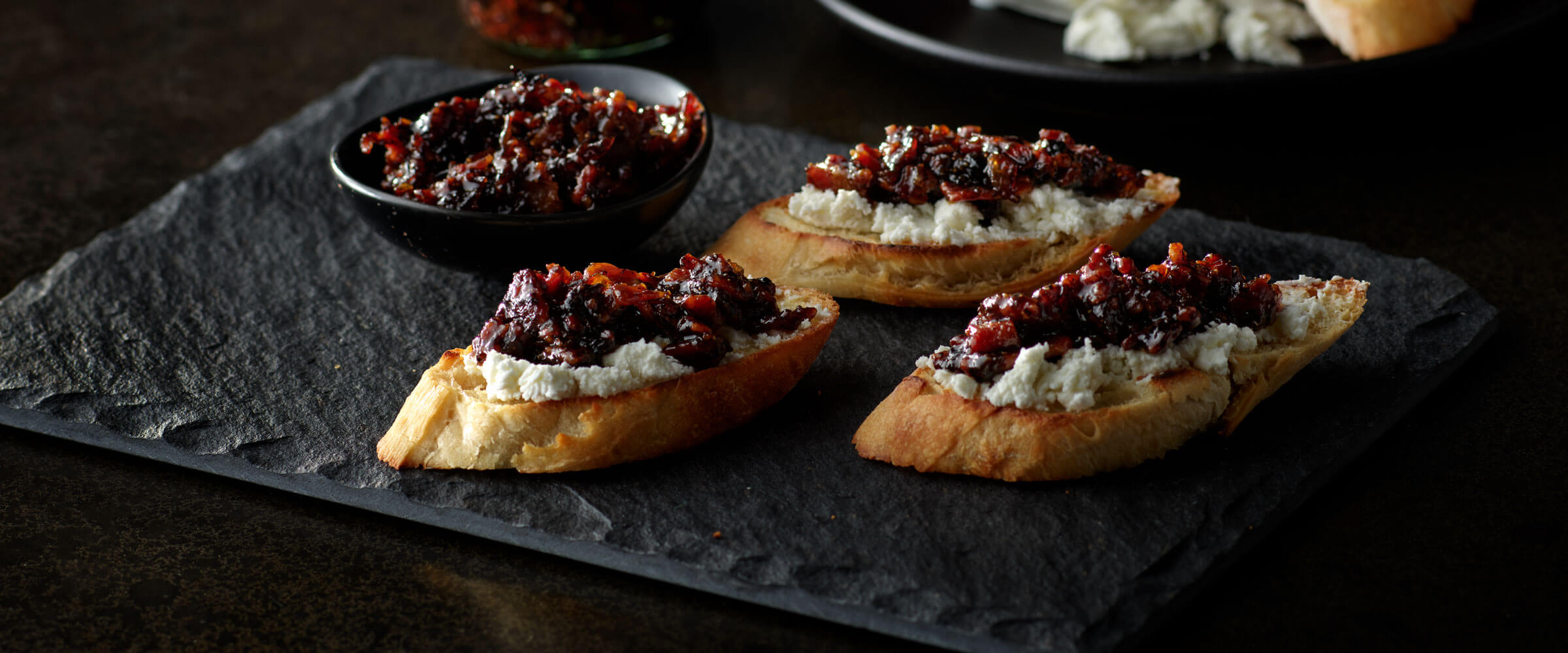 Bacon Jam on sliced baguette on black board with extra jam