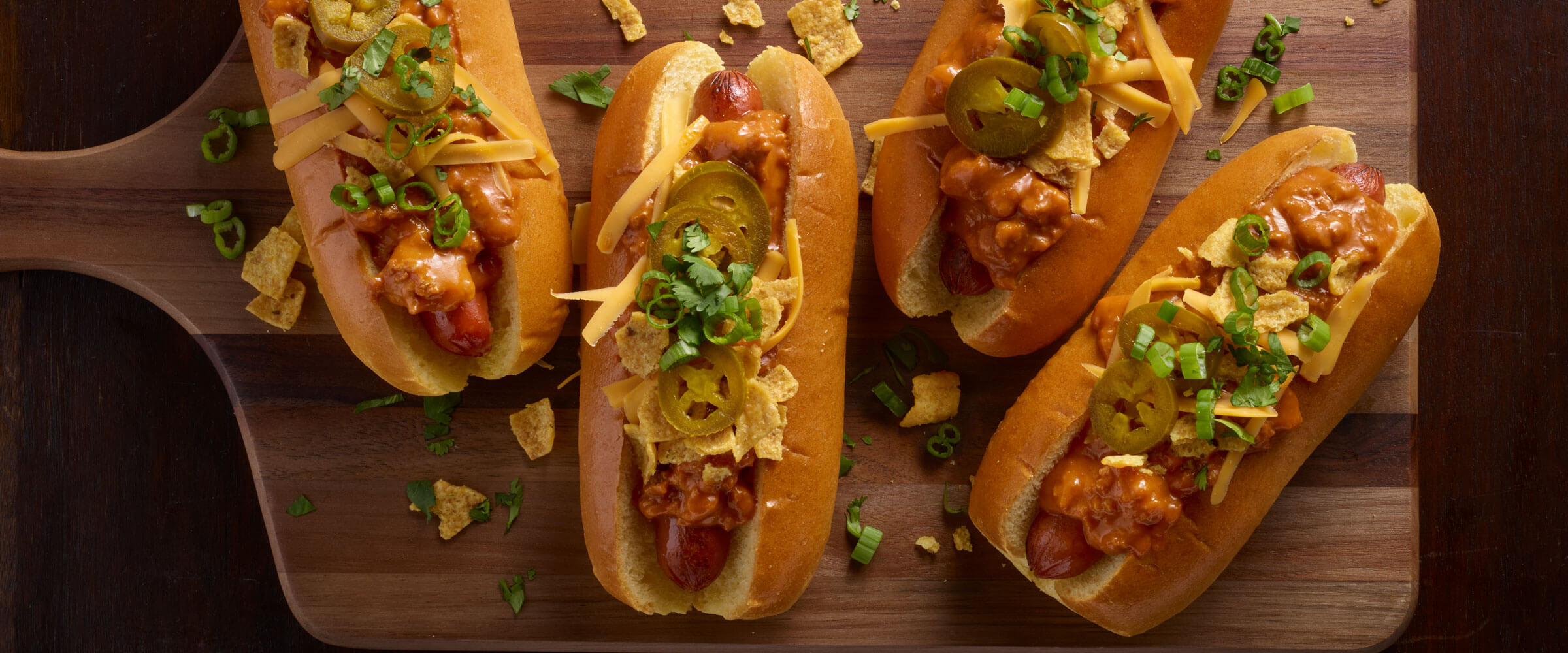 Cheese Chili Corn Chip Pie Chili Dogs topped with jalapenos, chips, and cilantro on wood board