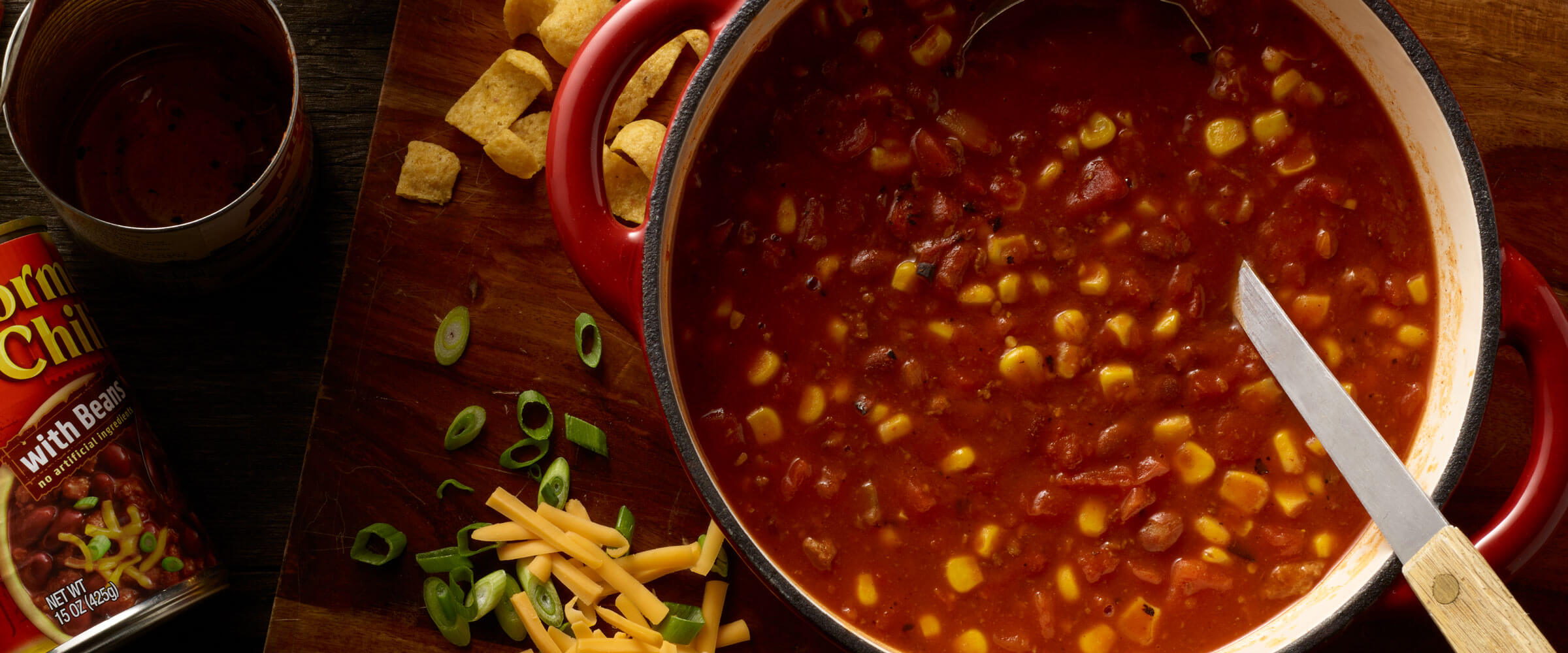 Chili with corn in red dish with serving spoon and toppings on the side