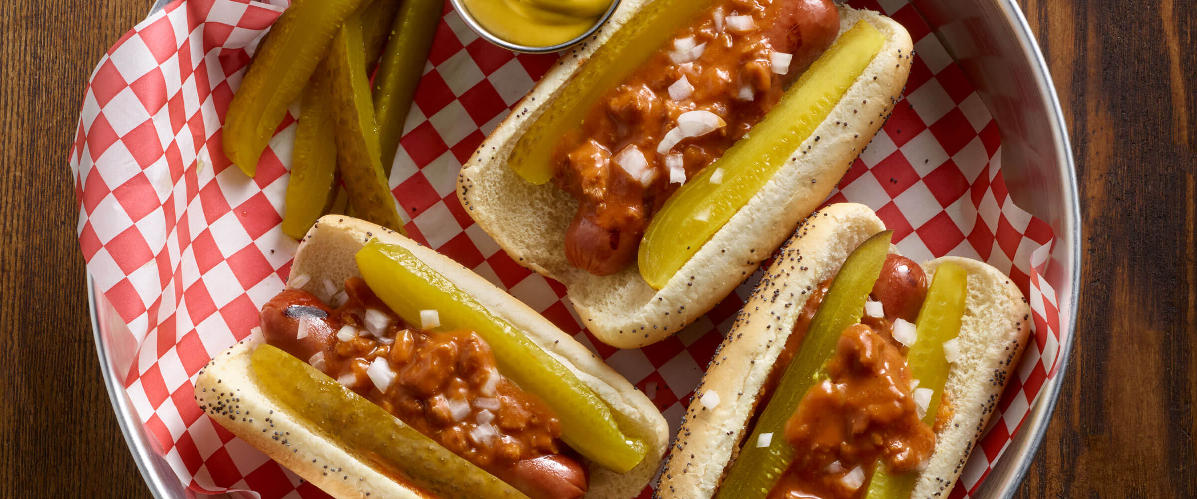 Chili Dilly Dogs topped with pickle slices and onion on red and white checked paper