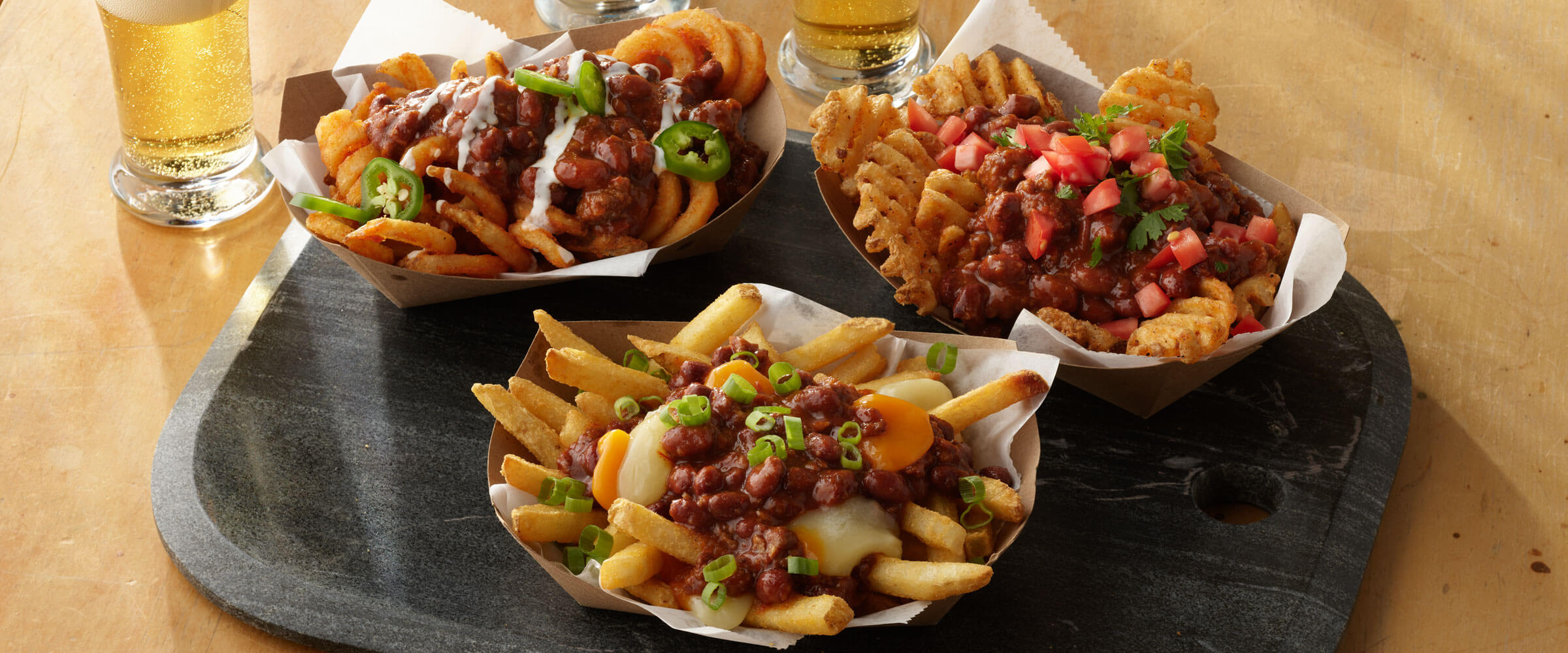 Chili Poutine 3 Ways topped with salsa, green onions and sauce in brown paper baskets with drinks on the side
