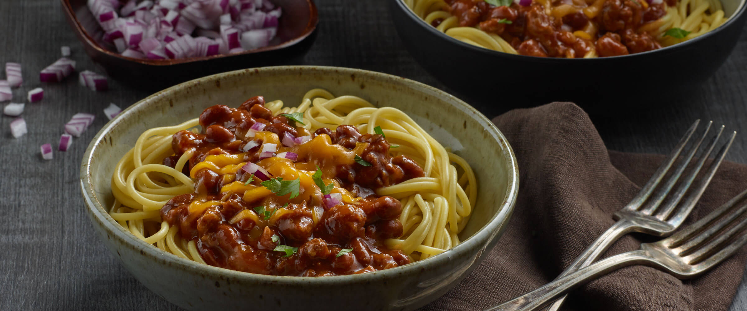 Cincinnati-Style Chili Spaghetti topped with cheese and red onions in dish with forks