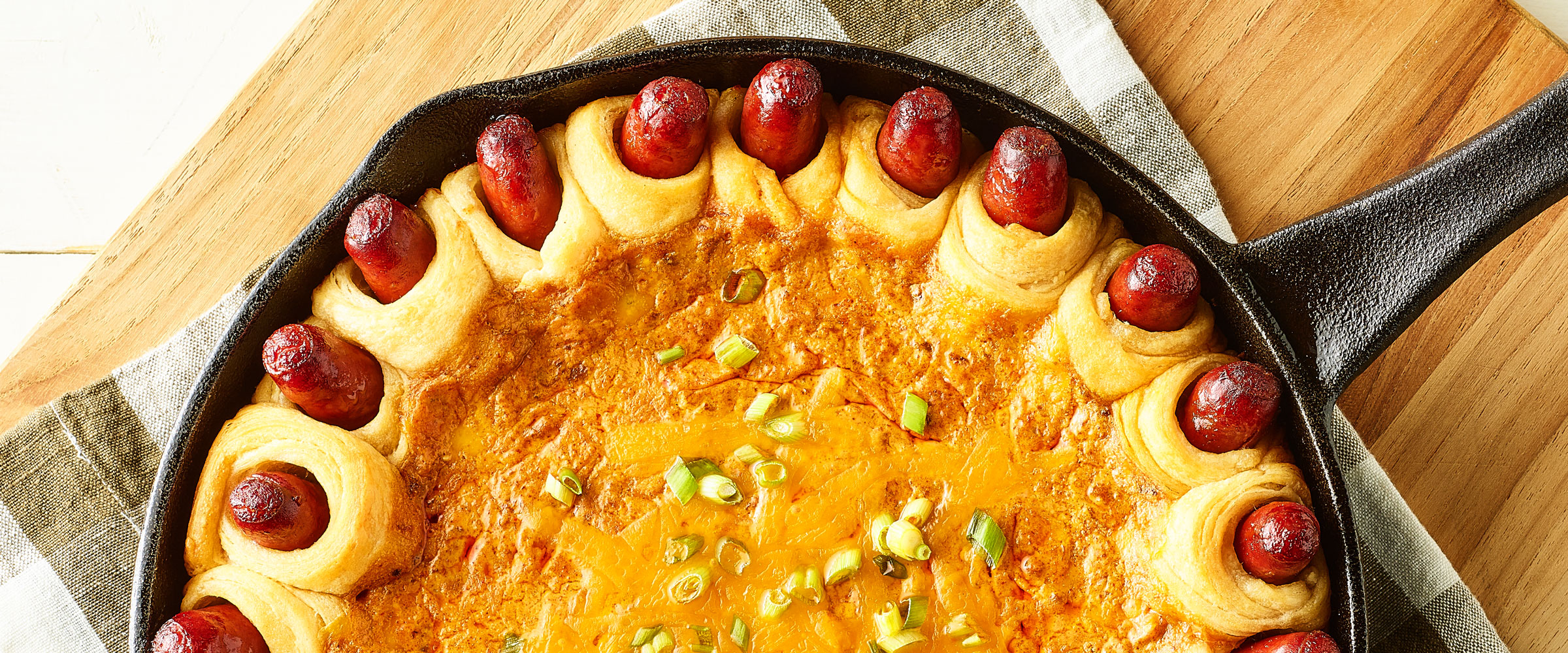 Chili Cheese dip in cast iron skillet with slices of hot dogs in pastry around the edge