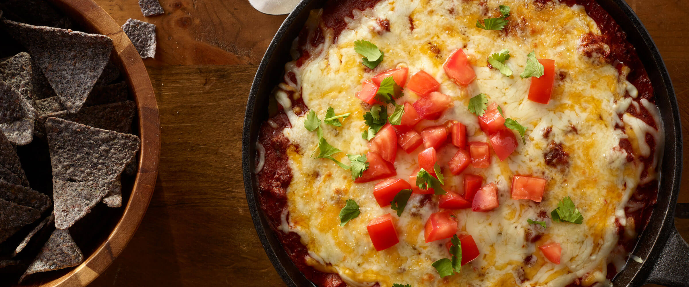 Layered Chili Cheese Dip in skillet topped with tomatoes with bowl of chips on the side