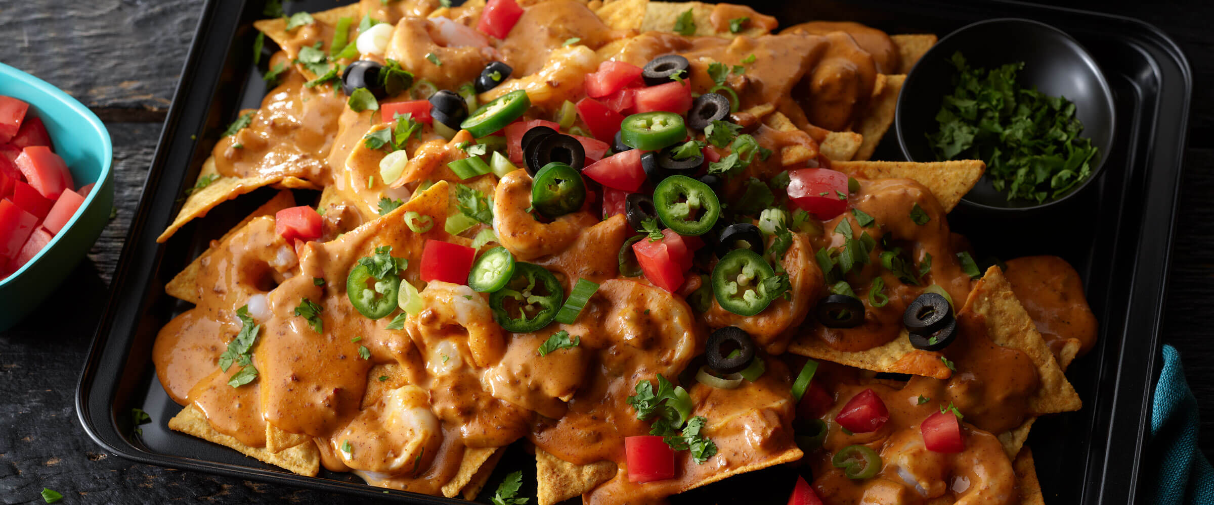 Sheet Pan Cajun Chili Shrimp Nachos in black dish topped with tomato, jalapeno, black olives and cilantro