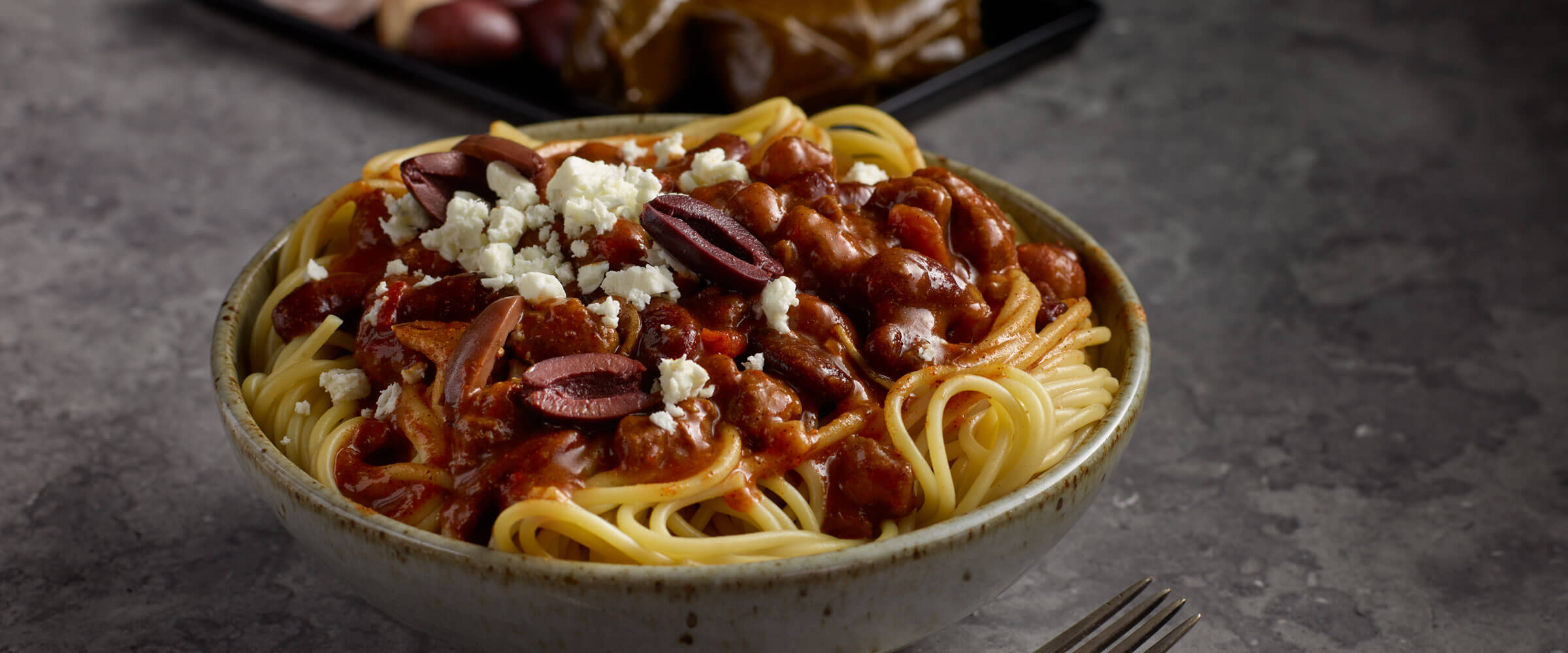 Big Greek chili spaghetti topped with olives in bowl on gray table