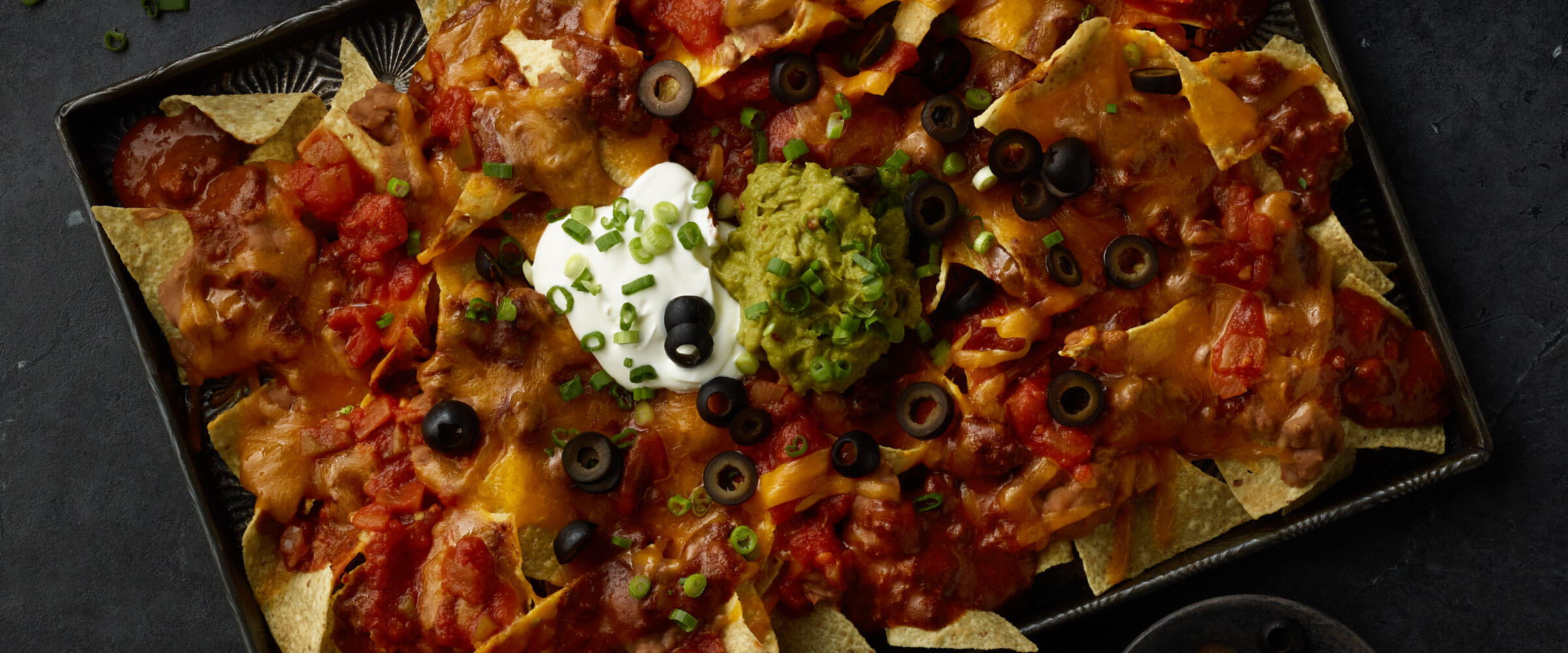 Ultimate Chili Nachos in black dish topped black olives, sour cream, chives and guacamole