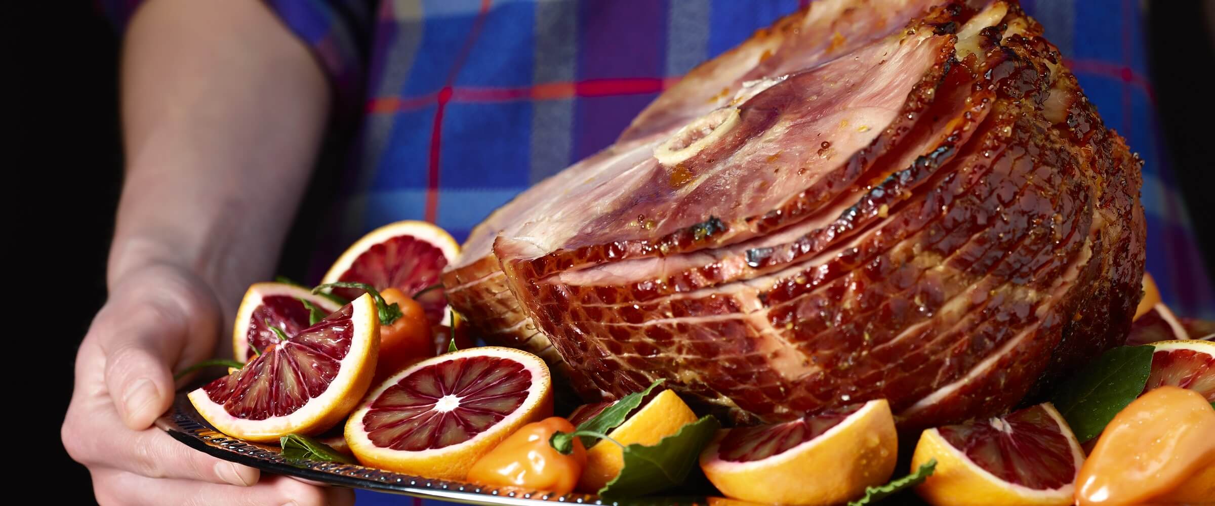 Person in blue plaid holding platter with sliced Blood orange habanero ham with orange wedges