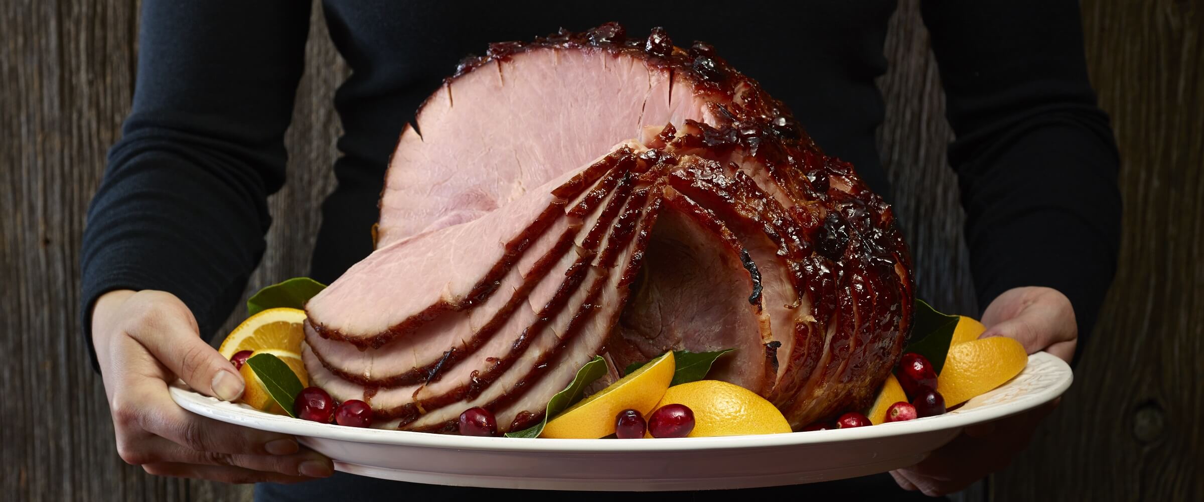 Person in black shirt hold platter with sliced Cranberry glazed ham with orange wedges