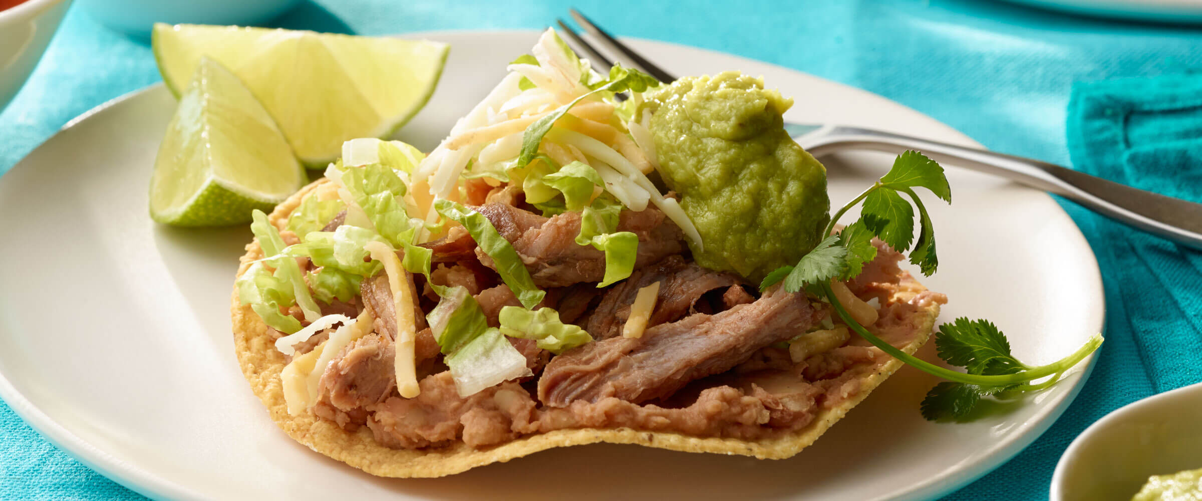 Southwestern Pork Tostadas topped with cheese, lettuce and guacamole on white plate with lime wedges