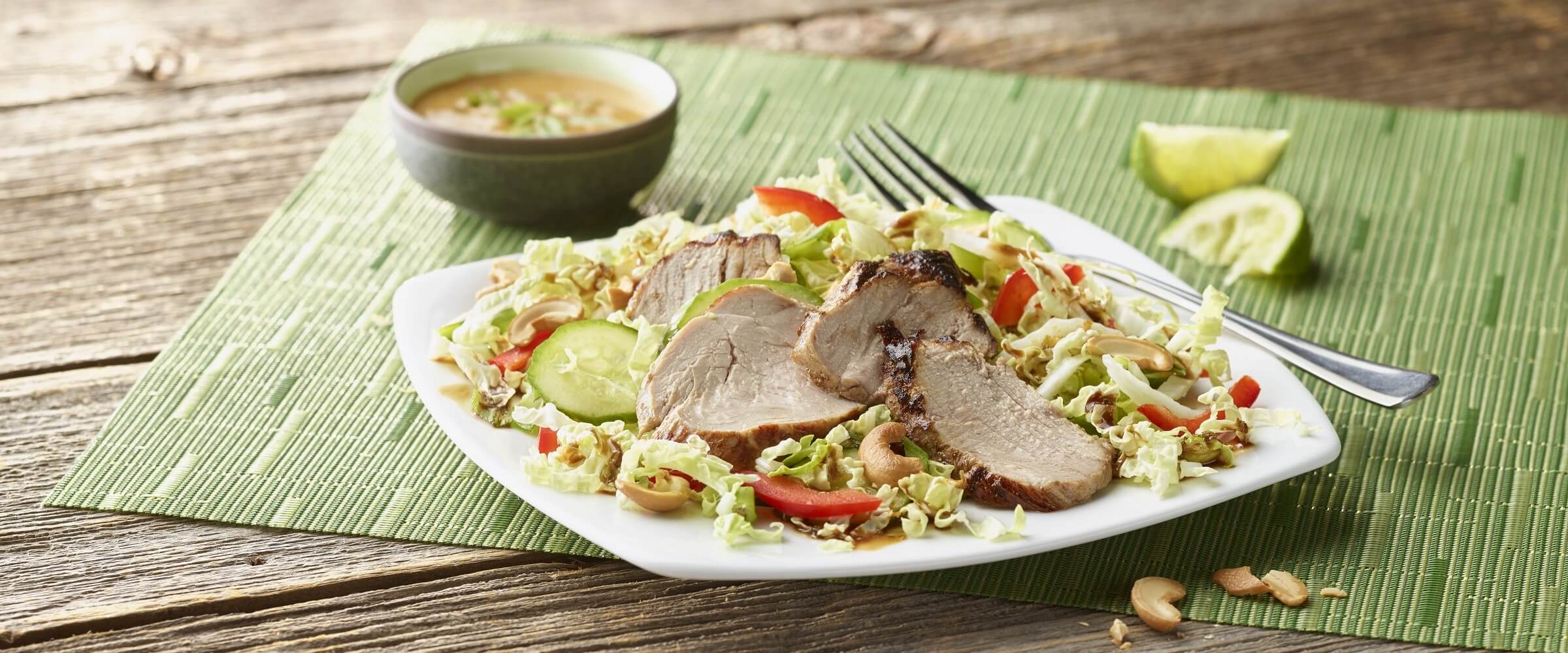 Thai pork tenderloin salad on white plate with dressing in bowl on green placemat