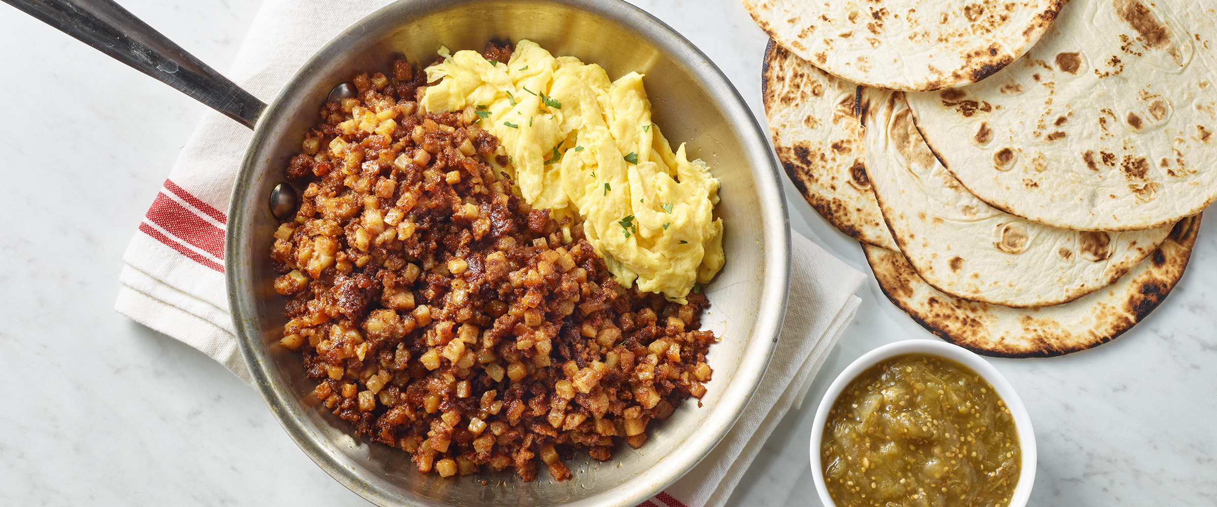 Chorizo and Eggs in a silver skillet. Next to the skillet is a small dish of salsa verde and tortillas.