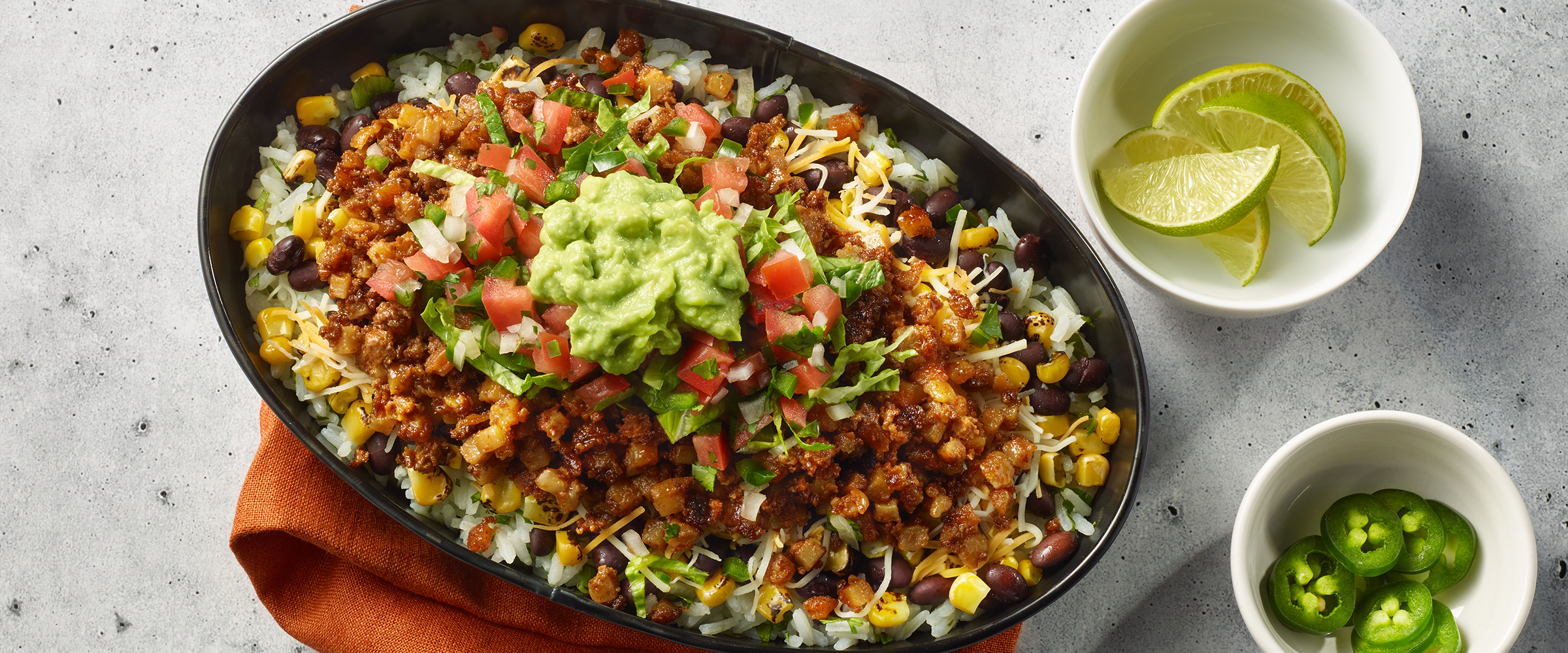 Chorizo Burrito Bowl in an oval cast-iron dish. Next to the dish is a small bowl of chopped green onions and wedges of lime.