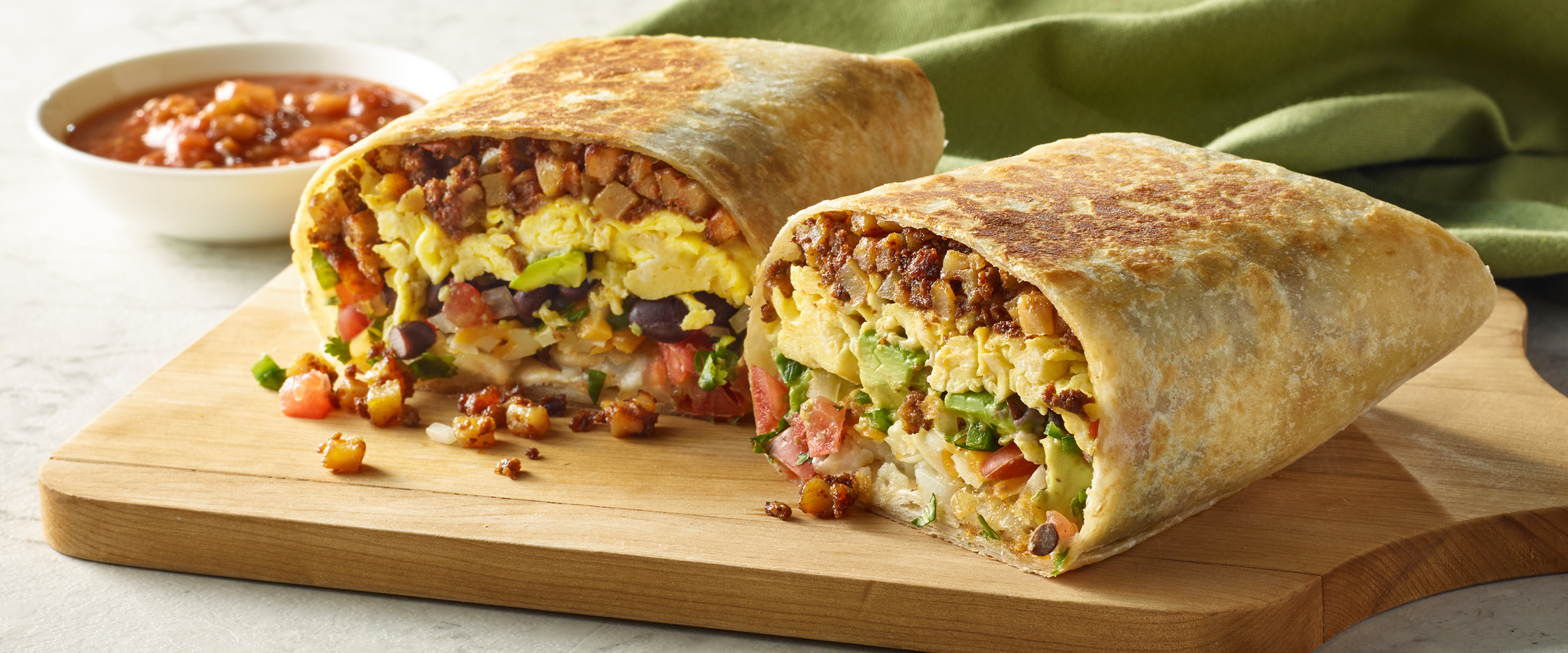 A Southwest Loaded Burrito cut in half on a wooden cutting board. Next to the cutting board is a small dish of salsa.