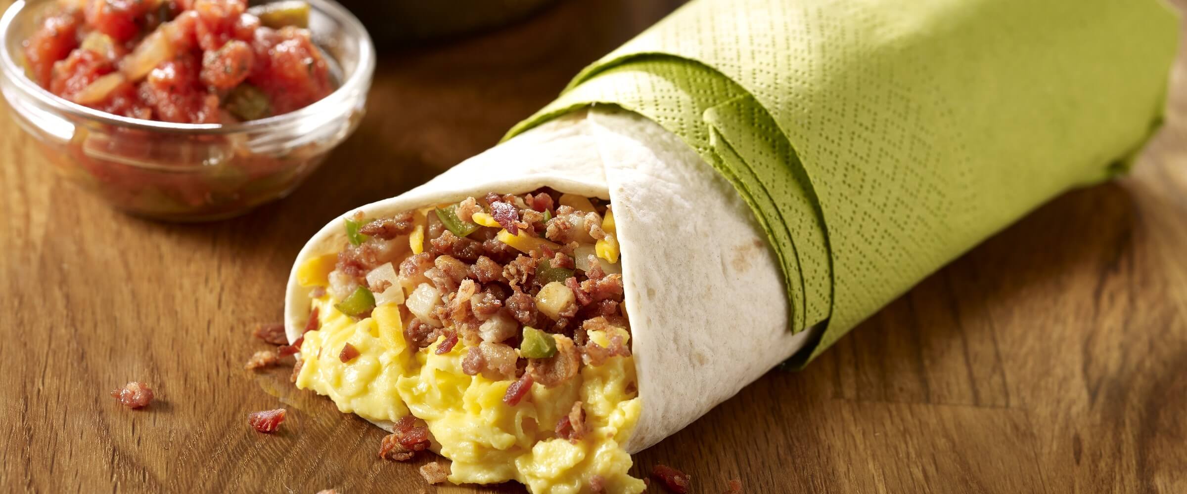 Corned beef hash egg burrito wrapped in green napkin with bowl of salsa