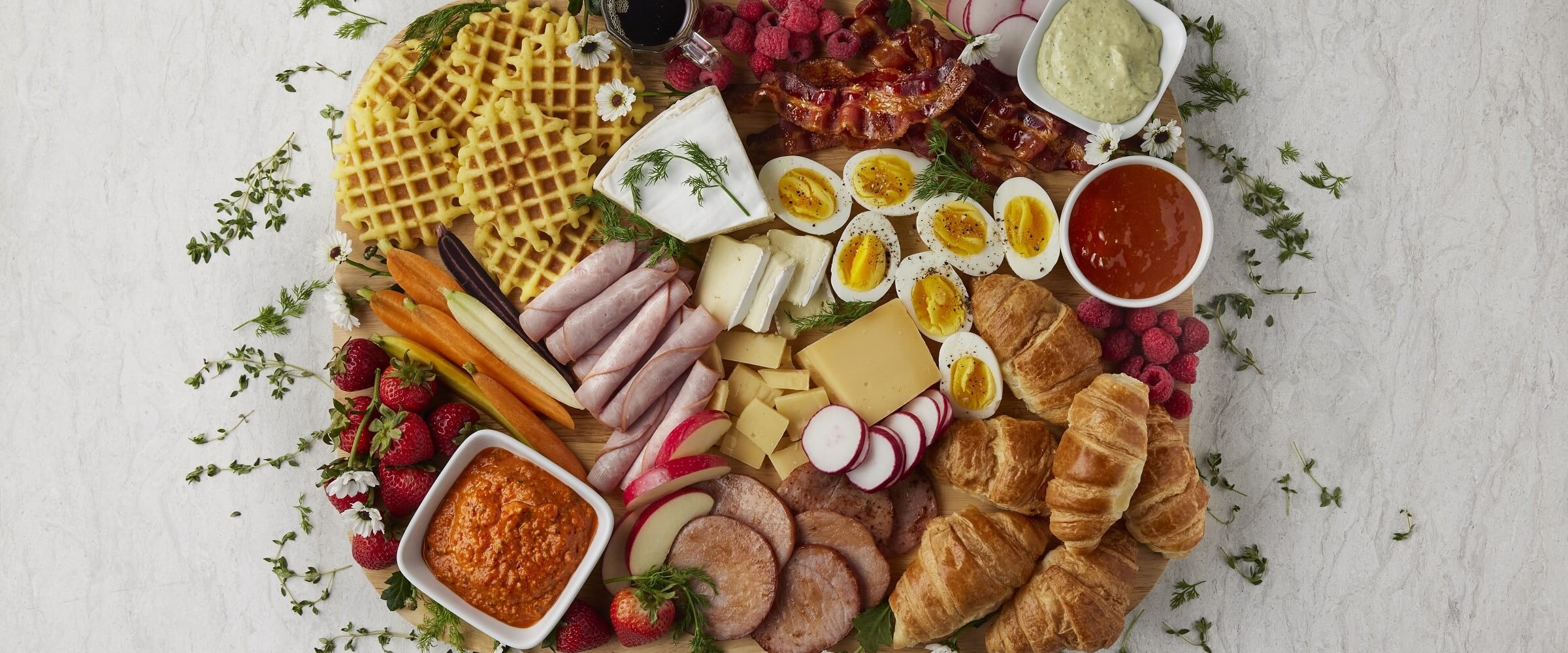 brunch board with meat, cheese, eggs, croissants, fruit, waffles, bacon and dip