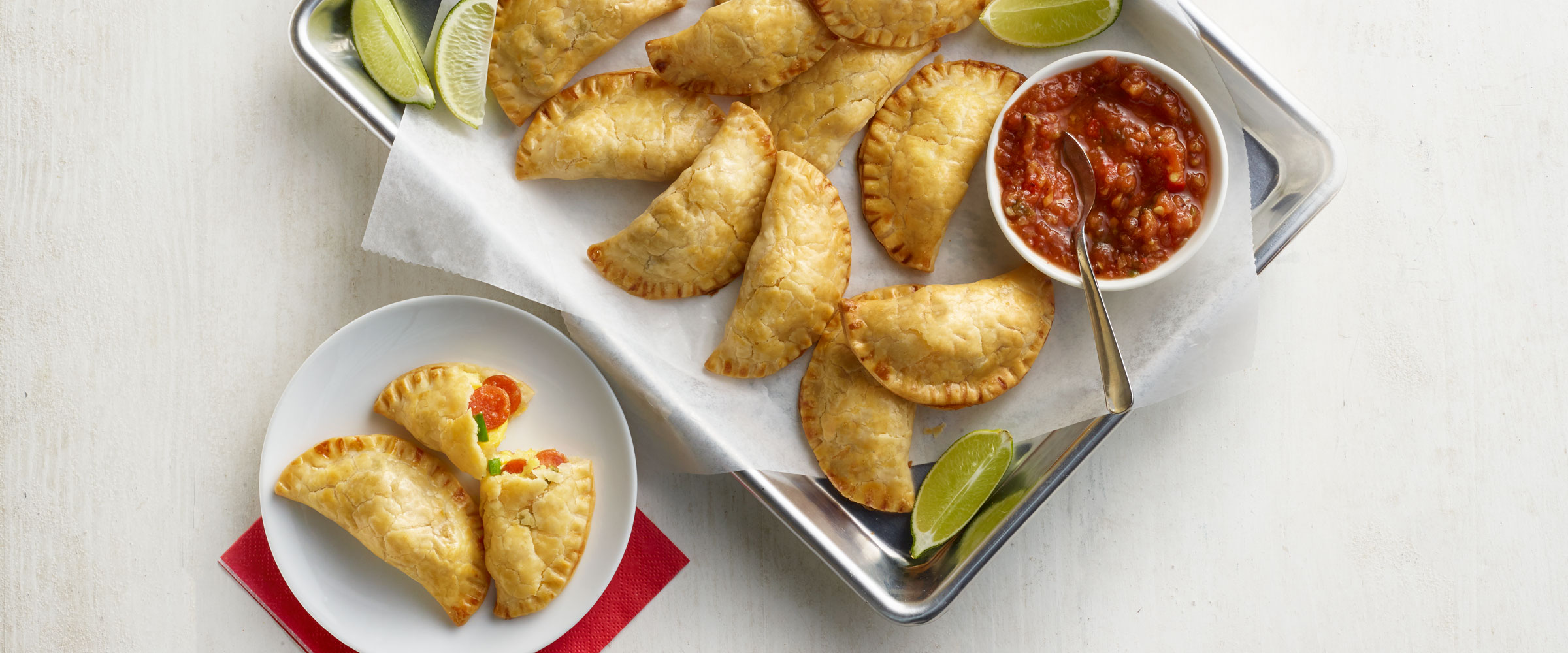 Pepperoni Breakfast Empanadas on sheet pan with dipping sauce and lime wedges