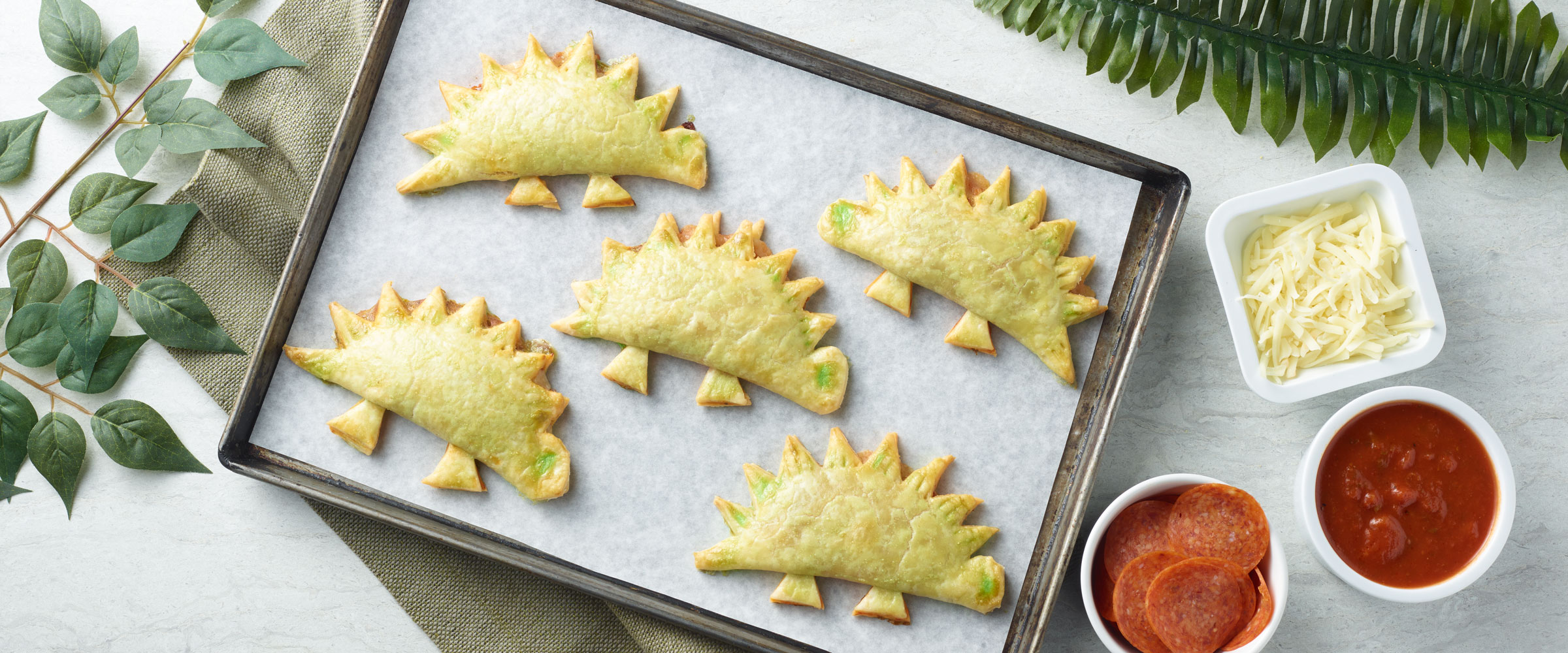 Pepperoni Dinosaur shaped Turnovers on sheet pan with toppings on side