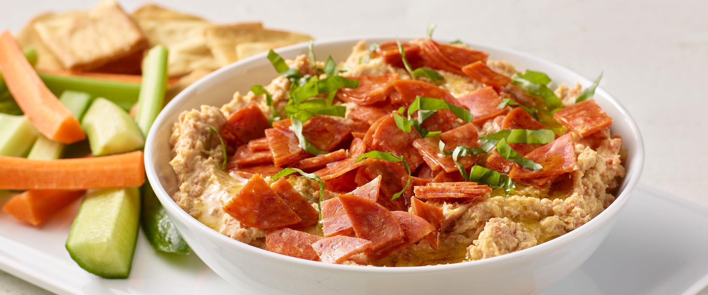 Pepperoni Hummus with garnish in white bowl with vegetables and crackers on side