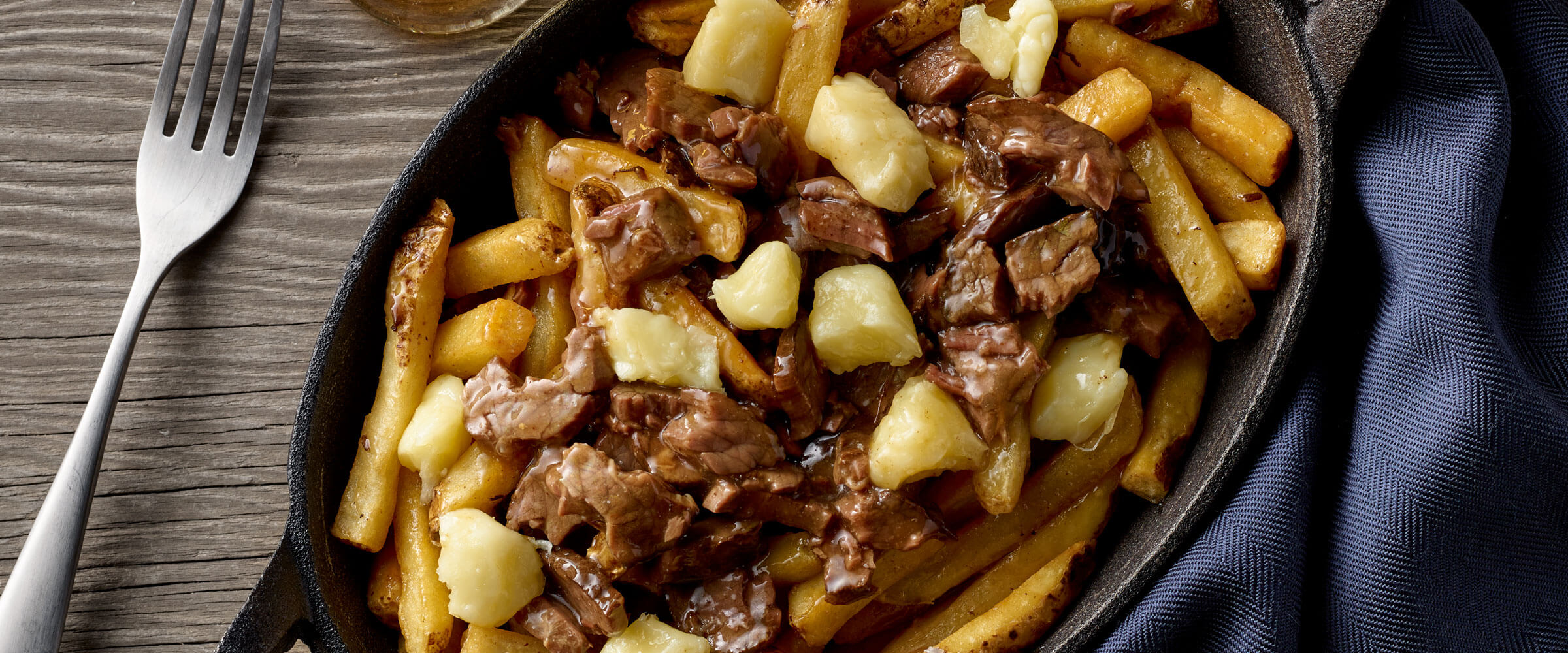 Roast Beef Poutine in cast iron skilled with blue napkin and fork