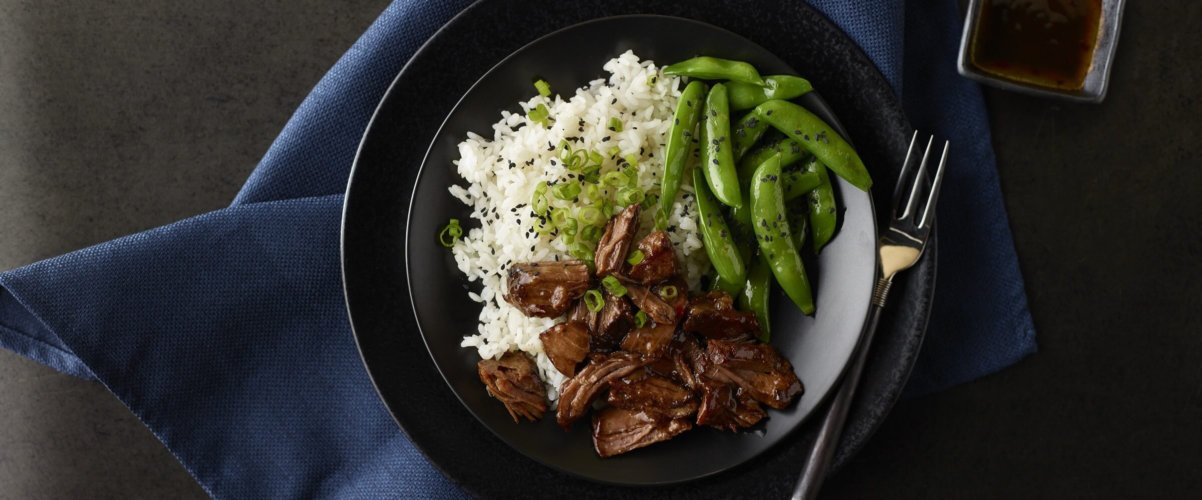 Sweet and sour beef with rice and pea pods in black bowl with blue napkin