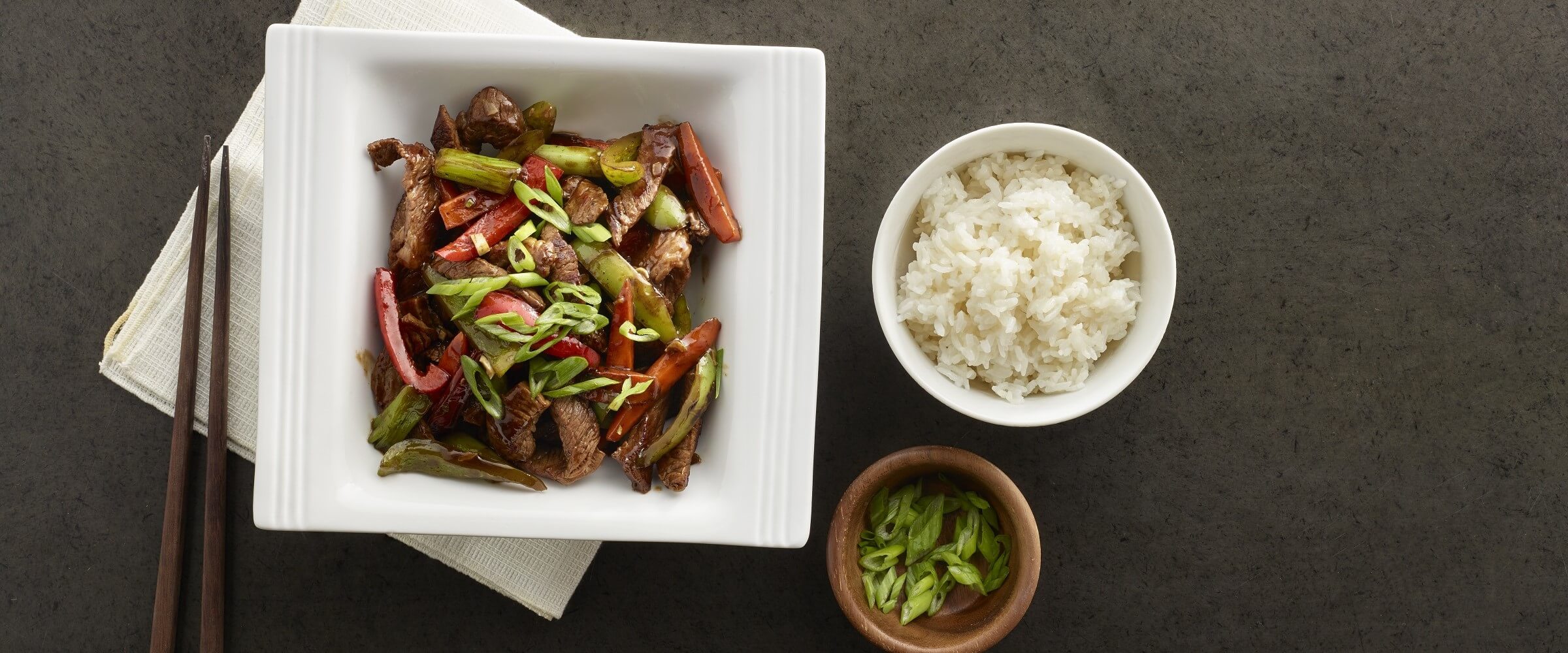 szechuan beef stir fry beef in white bowl with side of rice and green onions
