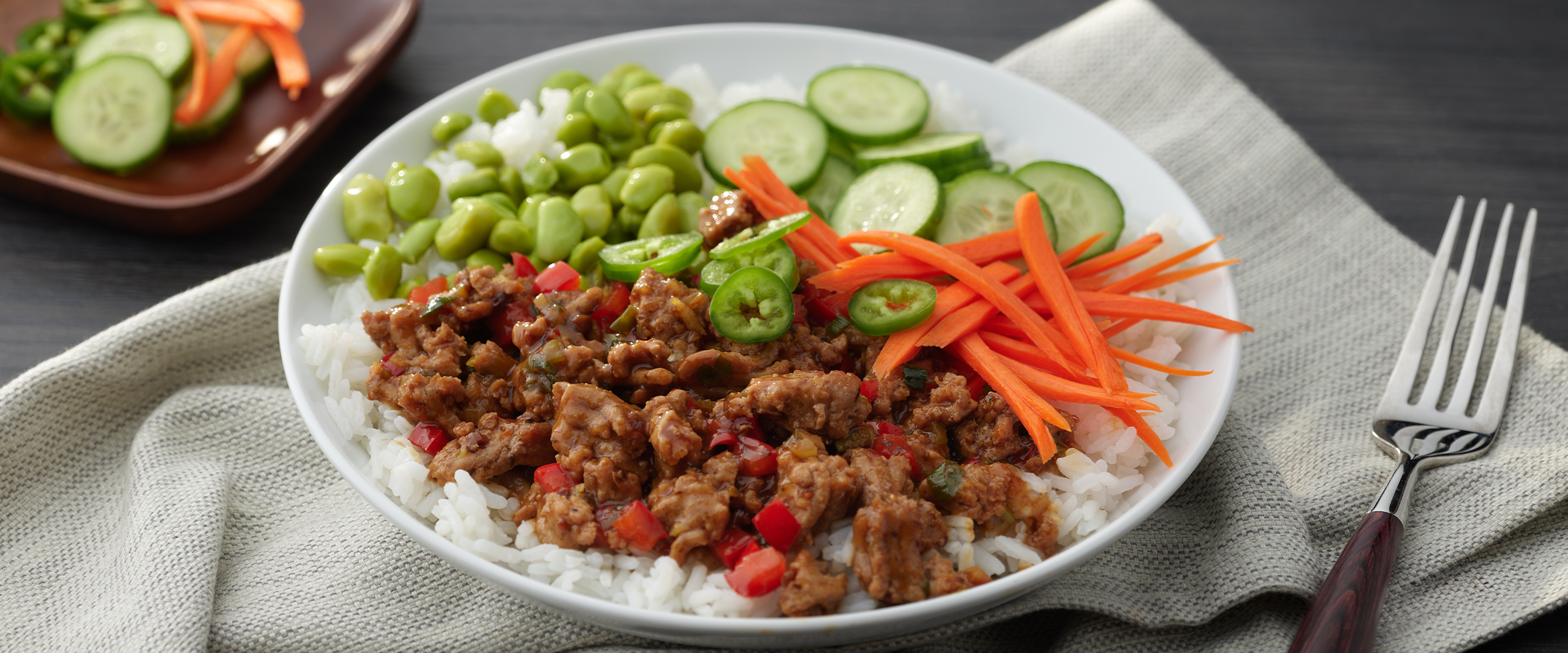 Teriyaki Turkey Bowl sitting on a white napkin. To the right of the bowl is a fork and to the left is a small bowl of sliced cucumber and julienned carrots.