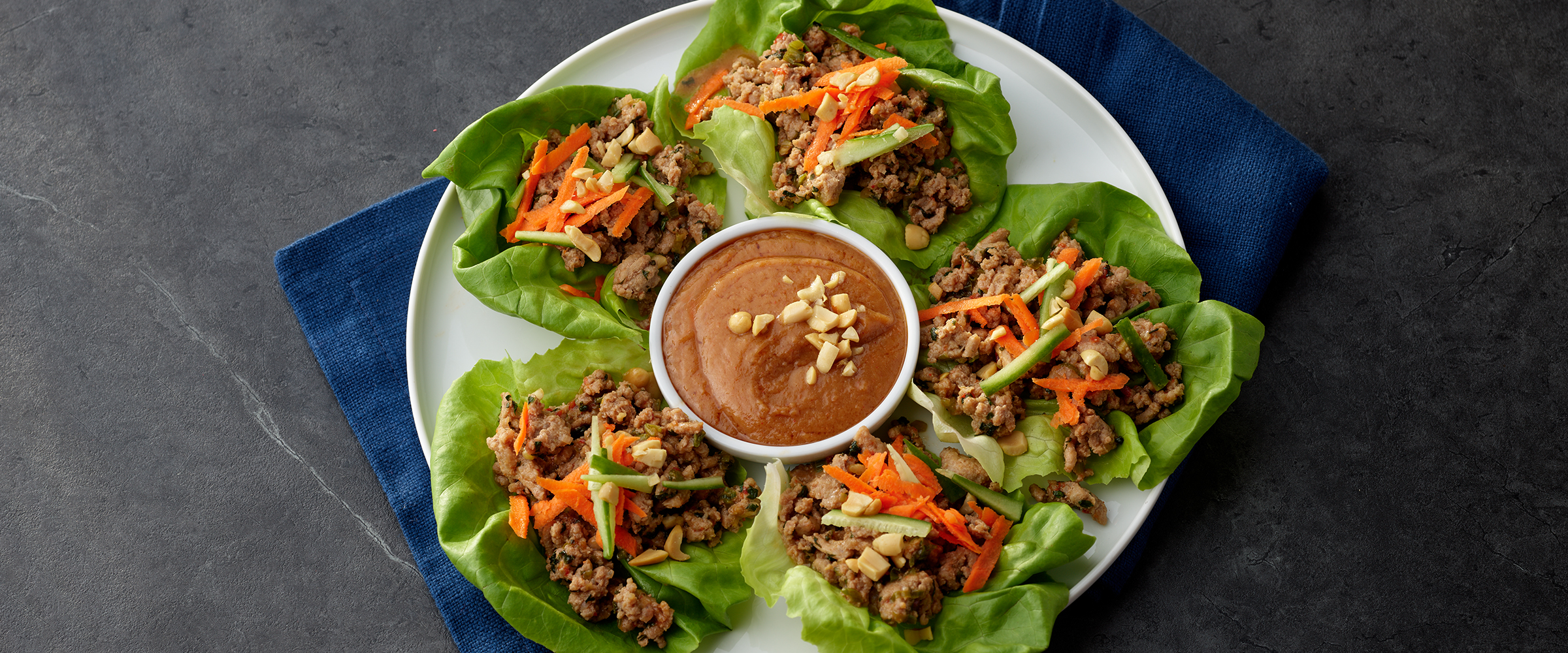 Five Thai Turkey Lettuce Wraps on a white plate. In the middle of a plate is a small dish with peanut sauce in it.