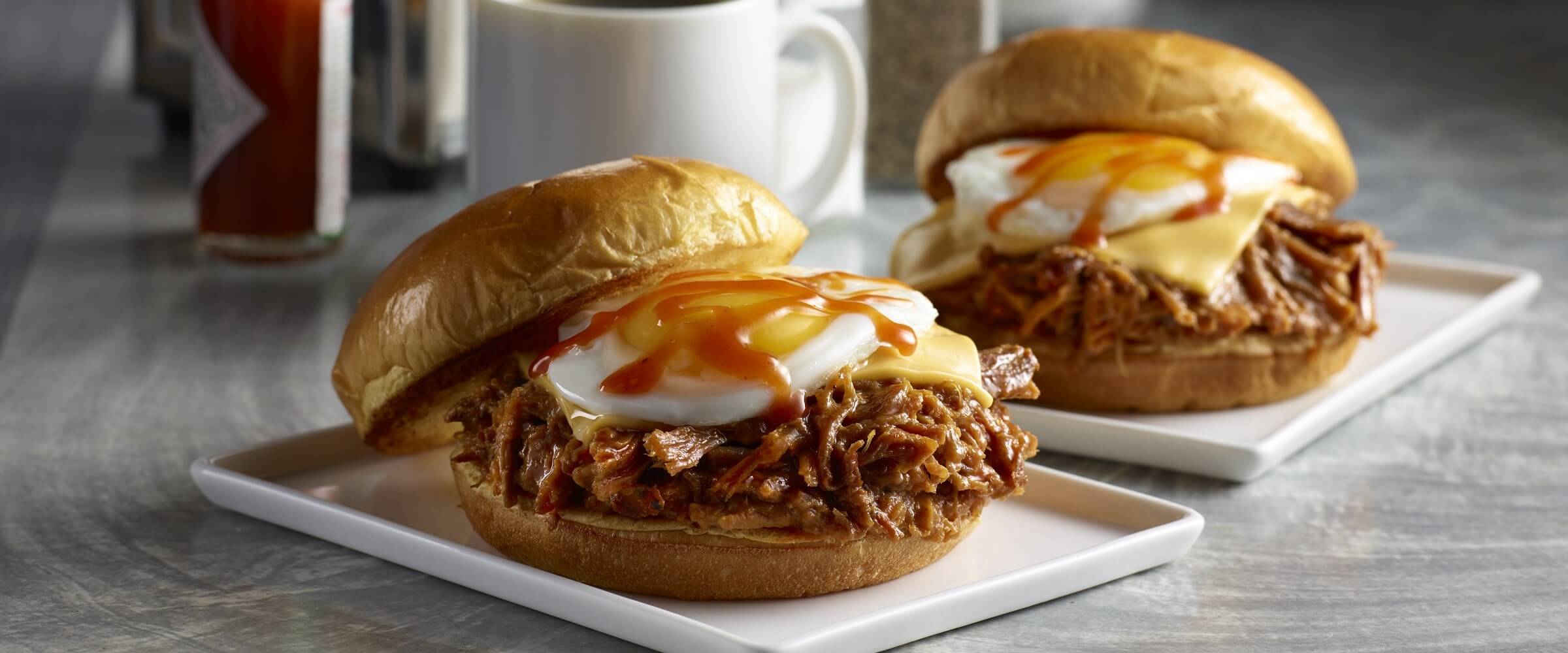 Pulled pork fried egg breakfast sandwich with cheese on white plates with coffee cup