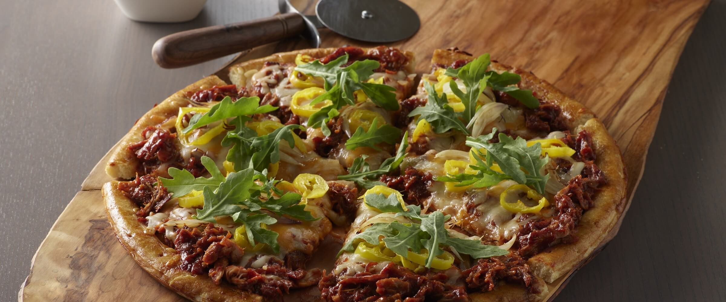 BBQ chicken pizza with arugula, shallot and banana peppers on wood cutting board with pizza cutter