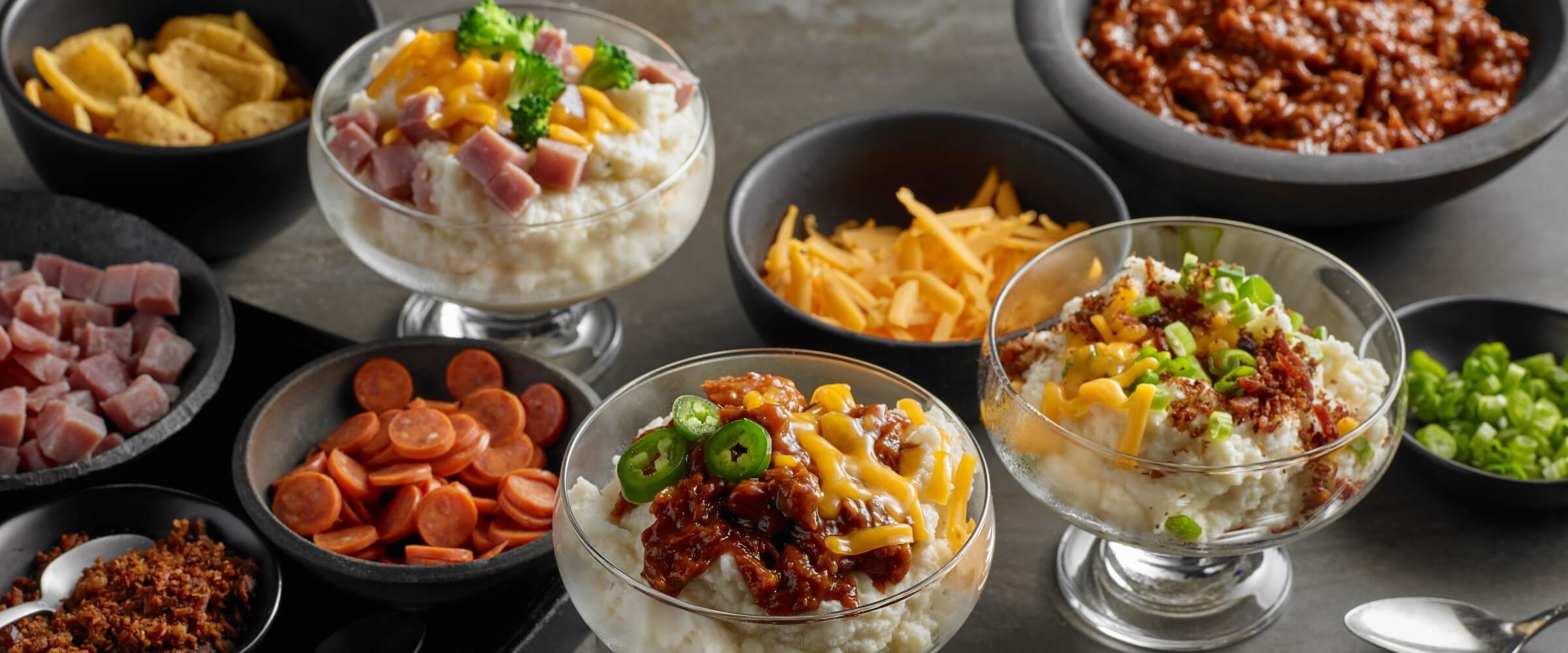 BBQ mashed potato bar spread with toppings in small bowls including pepperoni, cheese, ham, bacon bits and green onions