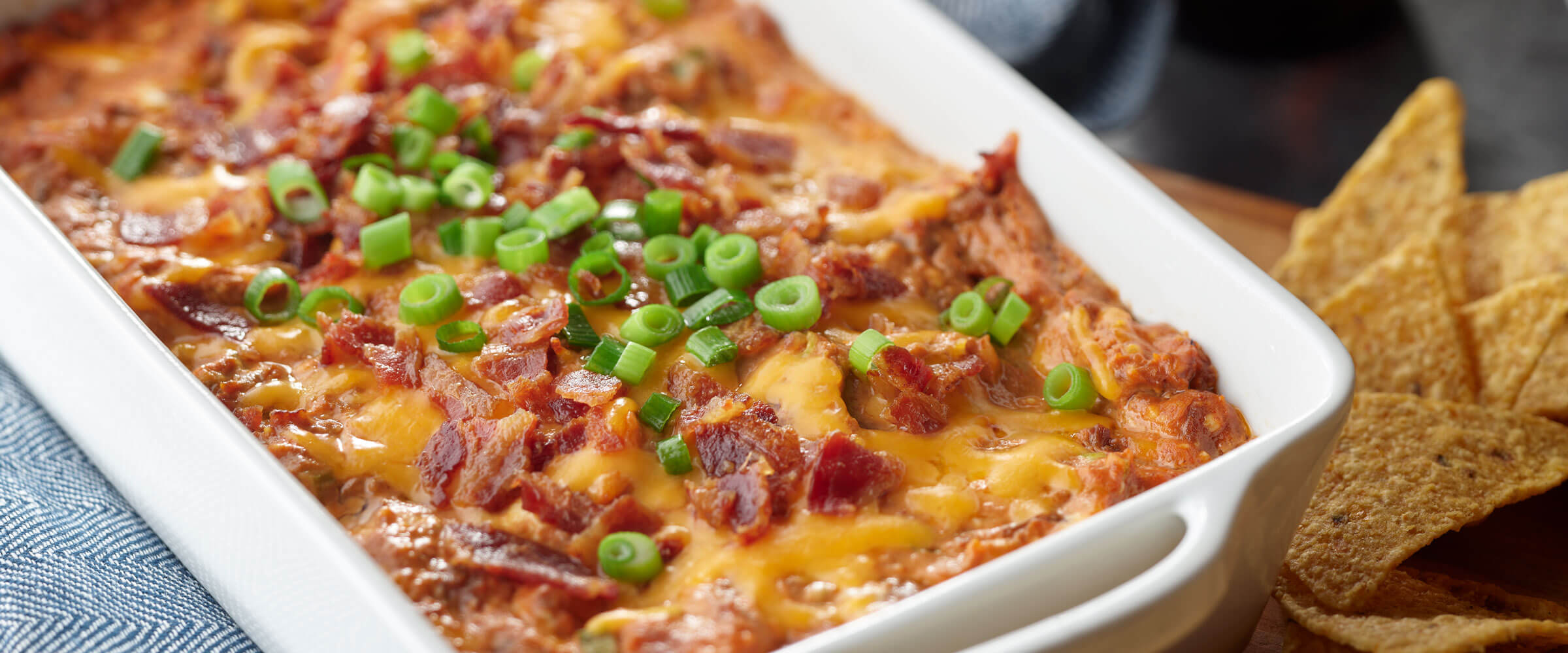 Cheesy Baked BBQ Dip in white dish topped with bacon and green onions with a side of chips