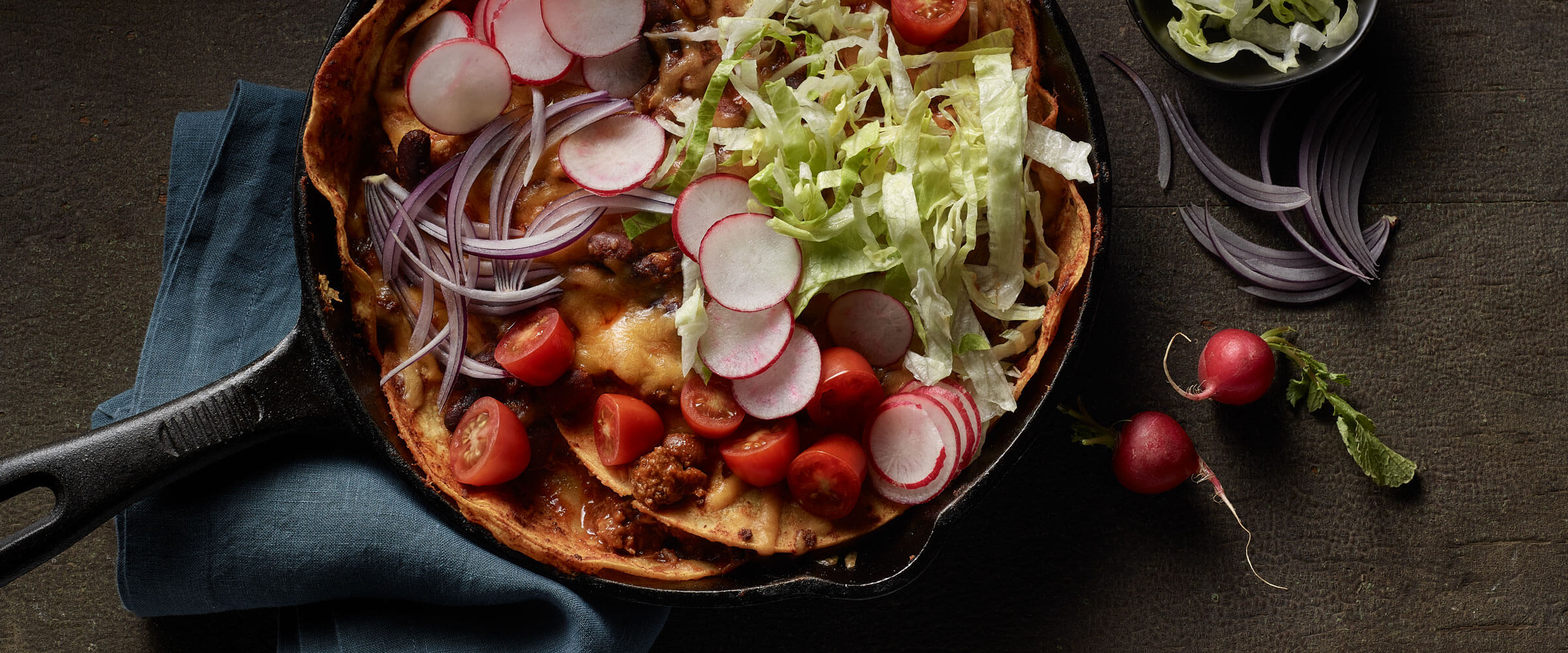 Layered Chili Enchiladas topped with radish, lettuce and tomatoes in skillet with blue napkin
