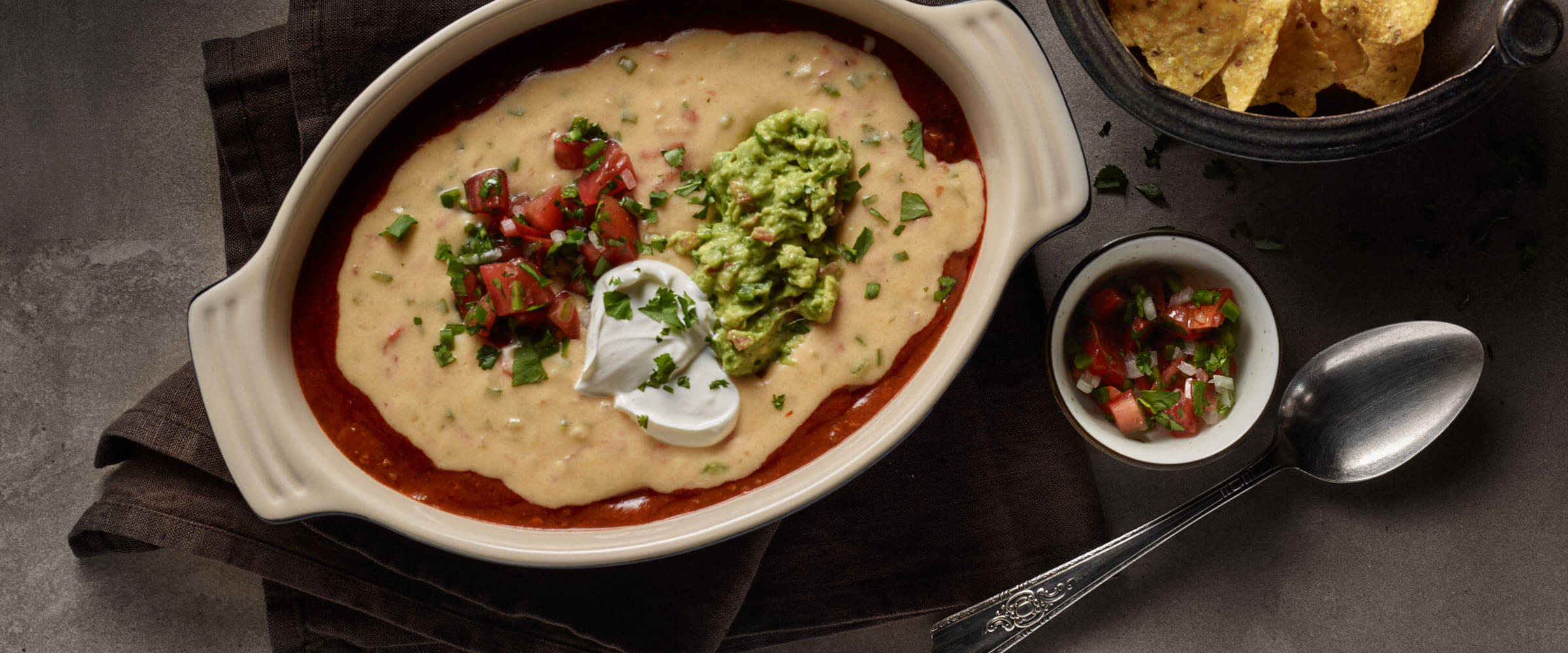 Chili Con Queso in white dish topped with salsa, sour cream and guacamole with side of chips