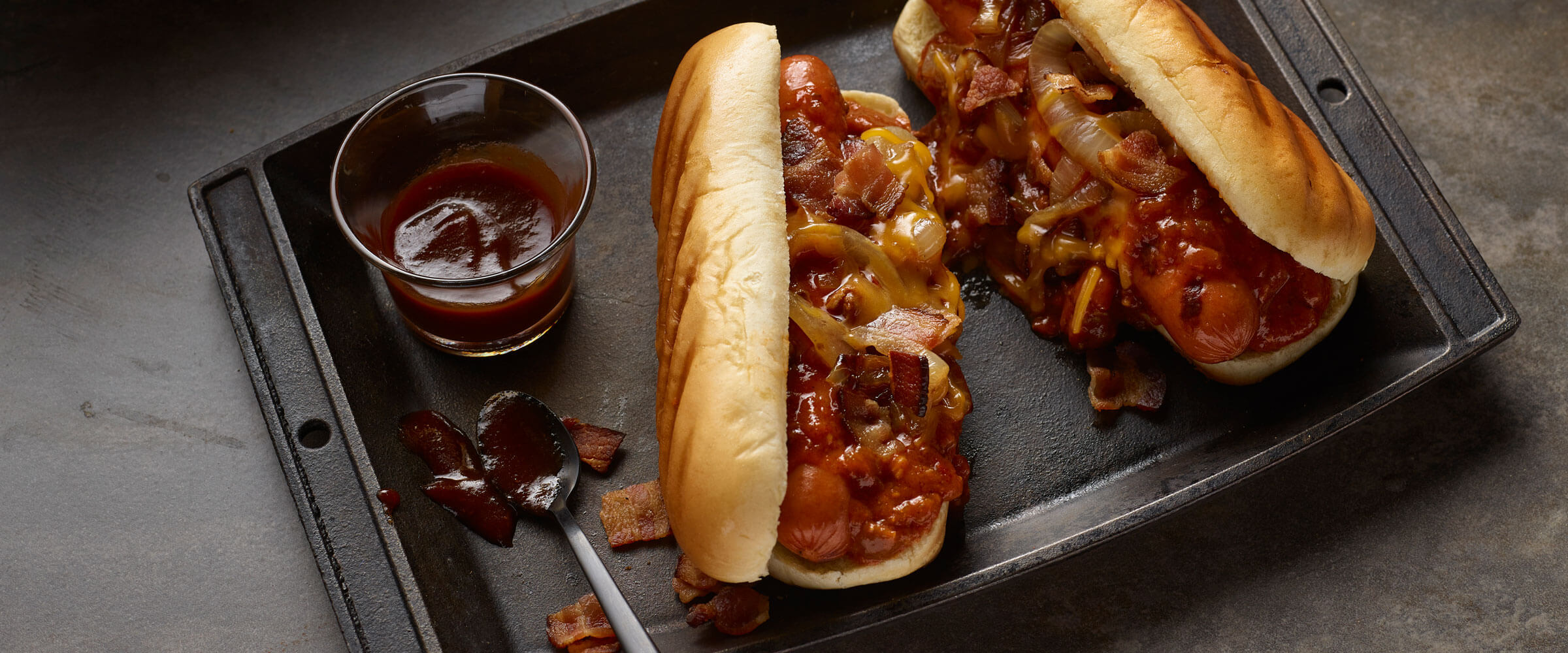 Cowboy Chili Dogs topped with onion and bacon with dipping sauce in baking pan