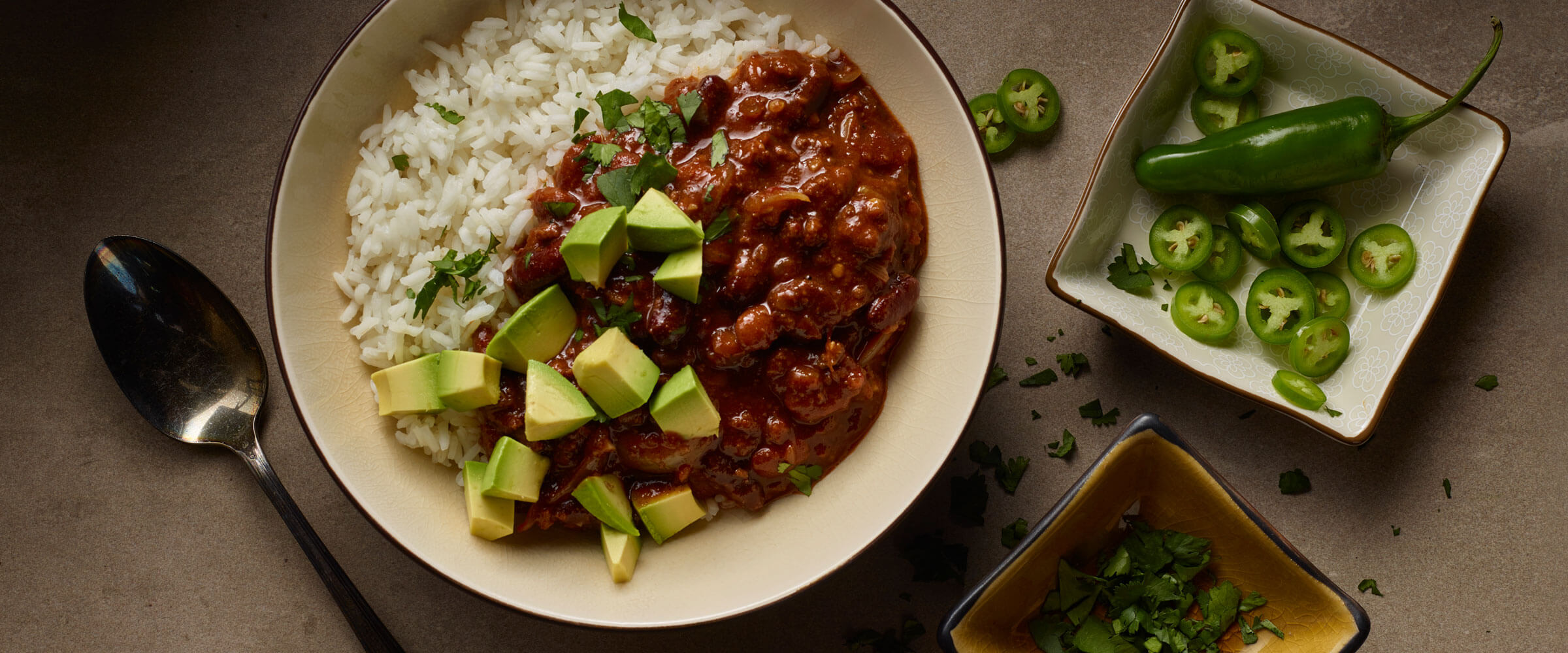 Bowl of chinese chili over rice topped with avocado and cilantro with dish of extra jalapeno slices
