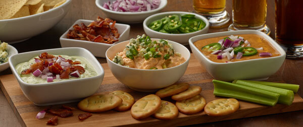 Spread of Game Day Dips in white bowls on wood board with crackers and celery