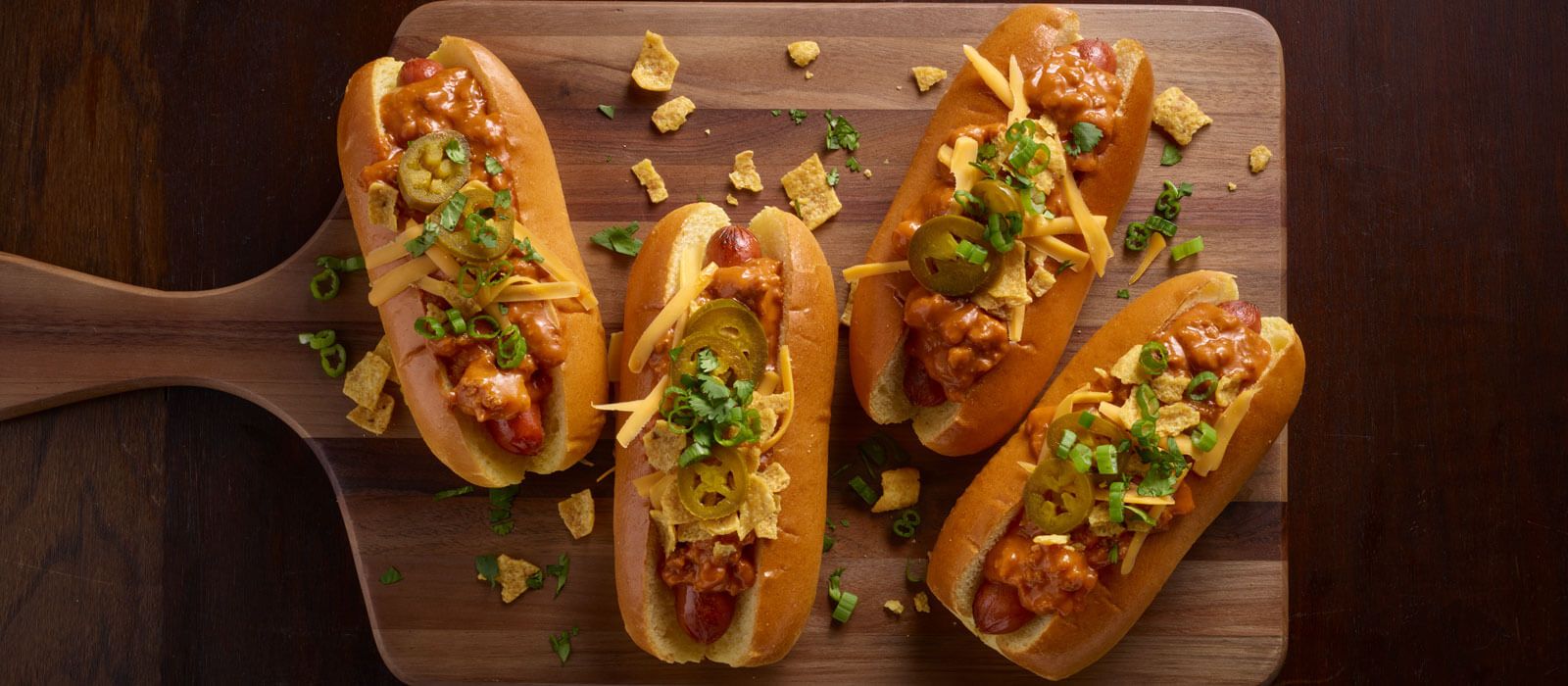 Hot dogs on wood cutting board topped with chili, jalapenos, cheese and chips