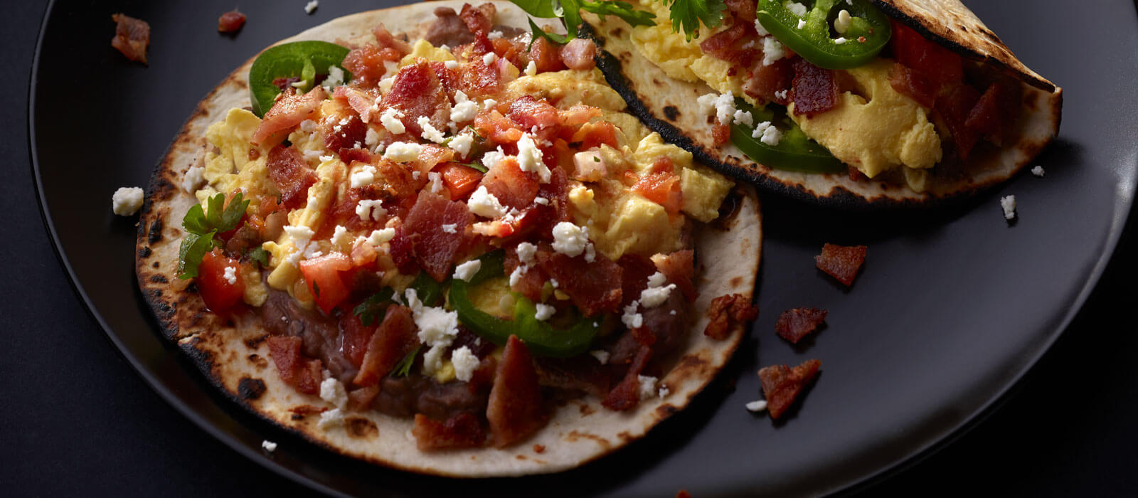Tortilla tacos topped with eggs, bacon, cheese and jalapenos on black plate
