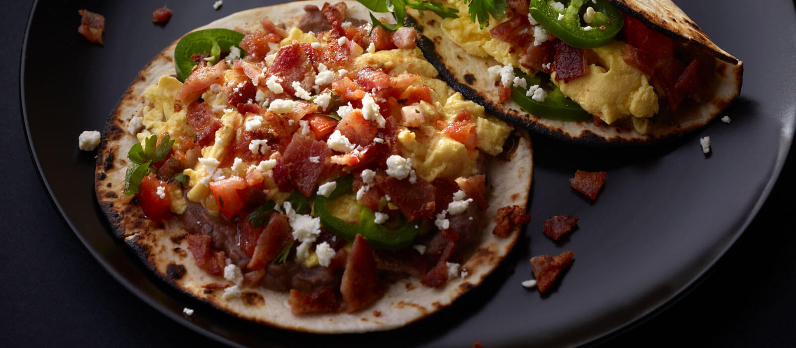 Tortilla tacos topped with eggs, bacon, cheese and jalapenos on black plate